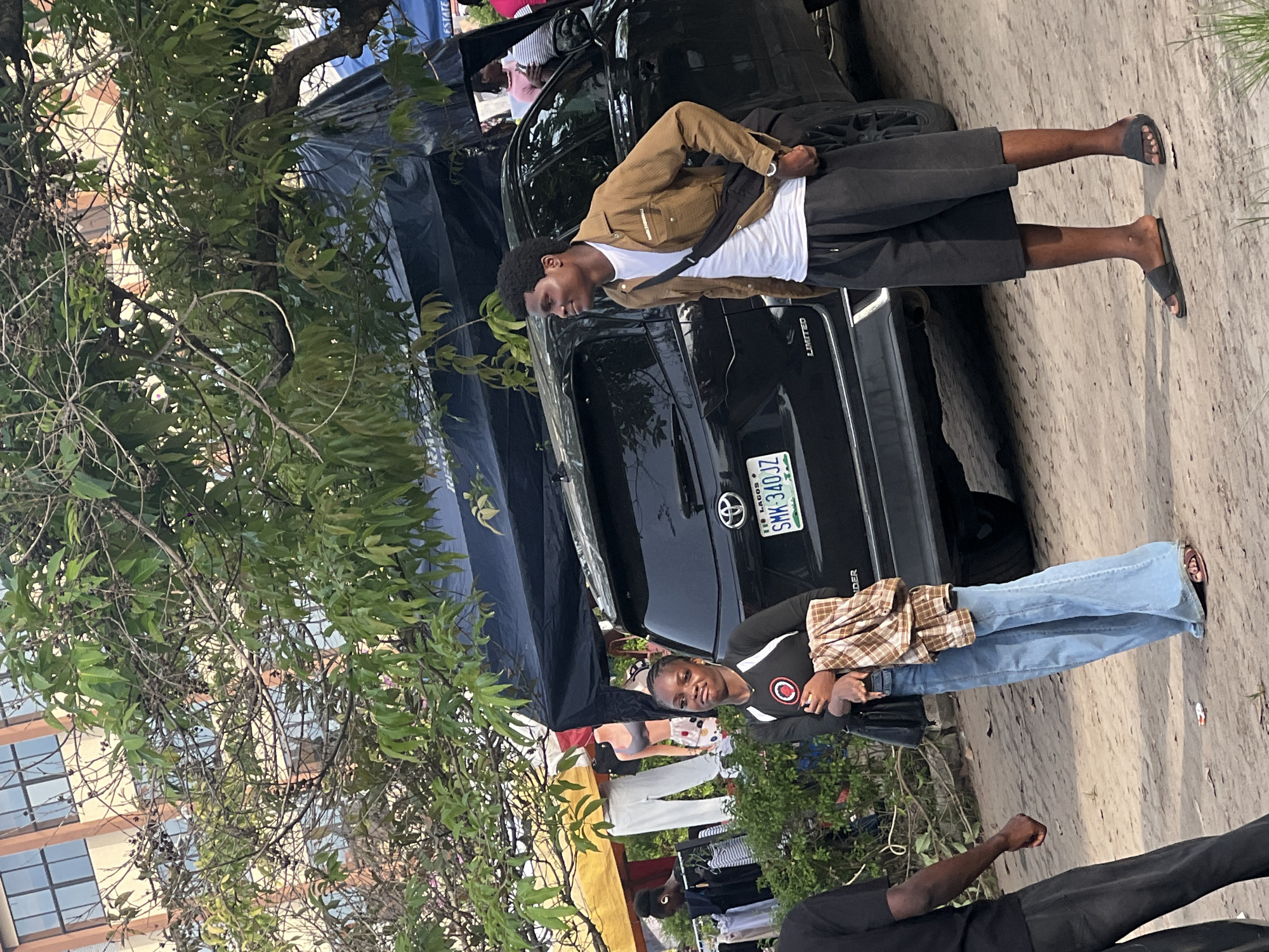 The image shows two people standing near a black SUV in Ijanikin, Nigeria. A man wearing a brown jacket, white shirt, and black shorts stands on the passenger side of the vehicle, with a black bag slung over his shoulder. On the driver's side, a woman in a black and white top and blue jeans leans against the car. The SUV has a Nigerian license plate (SMK-340JZ). Clothing racks with garments are visible in the background, suggesting a market or retail setting. A tree with dense foliage partially obscures the view of a building in the background. The ground appears to be paved or covered in dirt. A blue tarp is set up behind the car.