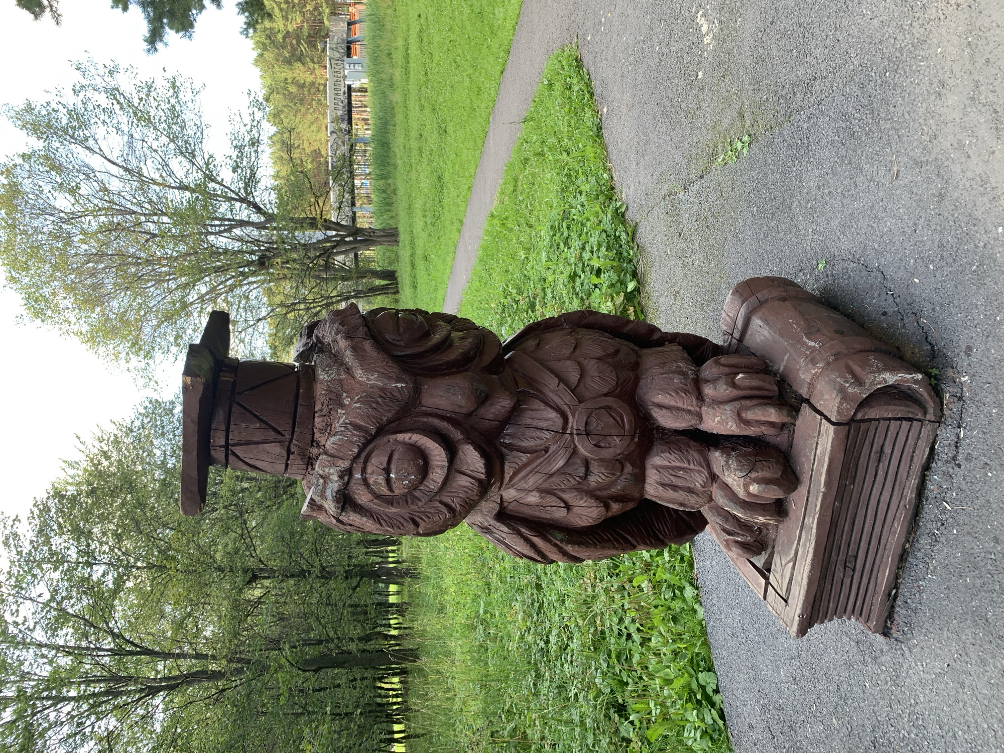 A carved wooden owl statue wearing a graduation cap stands on a book in a park. The owl is intricately carved, with detailed feathers and large, expressive eyes. The book it stands on is also made of wood and appears to be aged. The statue is placed on a paved pathway in a green park with trees in the background. In the distance, a bridge with text that reads "ПРОКОПЬЕВСКИ" (Prokopyevsk) is visible. The scene is set in the city of Prokopyevsk, Russia.