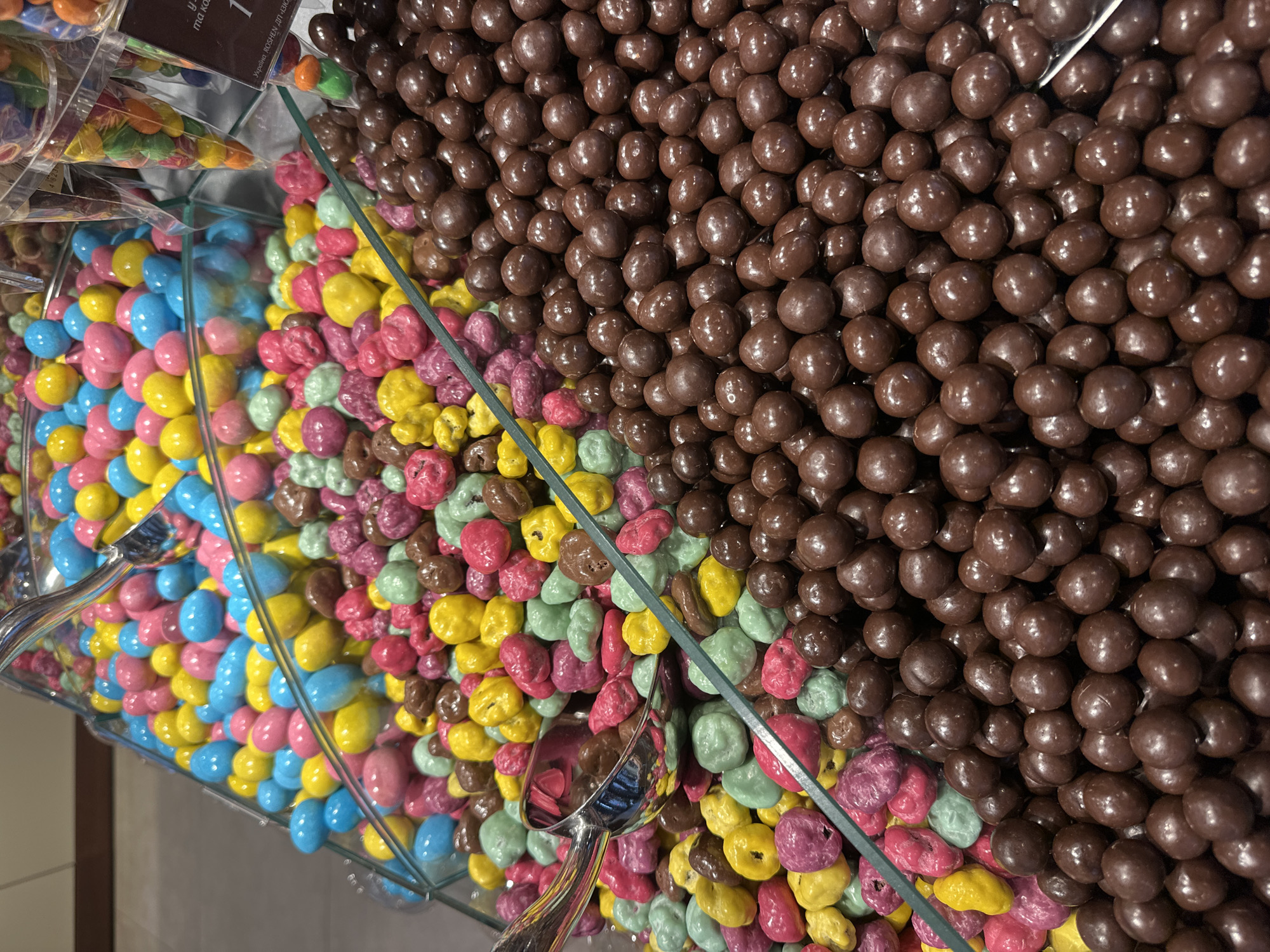 The image presents a detailed close-up of a bulk candy display within a retail environment, identified as being in Ternopil, Ukraine. The scene is brightly lit, showcasing various confectionery items organized in clear, compartmentalized dispensers.
In the upper left quadrant, a quantity of uniformly ovate candies with smooth, glossy coatings are visible, primarily in shades of bright blue, pink, and yellow. A metallic serving scoop is partially submerged within this selection. Moving to the lower-left and central areas, a second type of candy is displayed. These are irregularly shaped with textured, matte coatings, appearing in vibrant colors such as fuchsia, bright yellow, mint green, and dark reddish-brown. Another metallic scoop rests within this assortment.
The largest portion of the display, extending across the entire right side and bottom right, features numerous dark brown, uniformly spherical candies with smooth surfaces, resembling chocolate-coated nuts or malted milk balls.
In the upper right background, out of focus, several transparent plastic bags filled with smaller, multi-colored candies are visible, hanging from a rack. Below these bags, a white rectangular label with black Cyrillic text is partially discernible. The legible portion reads "Україна ROSHEN ДП «ДКК «", clearly identifying the Ukrainian confectionery corporation Roshen, thereby corroborating the specified location in Ukraine. The general background is indistinct, showing blurred warm tones and a lighter floor, consistent with an indoor commercial setting. No individuals are present in the frame.