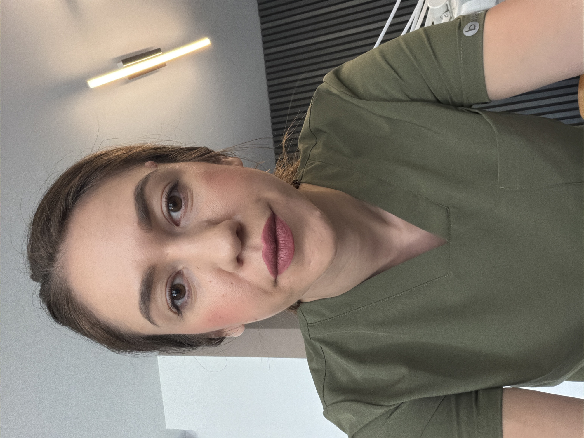 A woman is taking a selfie in what appears to be a professional setting. She is wearing a green scrub top, and her hair is pulled back. She has makeup on, including lipstick and blush. In the background, there is a modern light fixture on the ceiling and a wall with vertical lines. The overall impression is a clean, well-lit environment. There is no visual information to suggest the location is in the city of Sofia, Bulgaria.