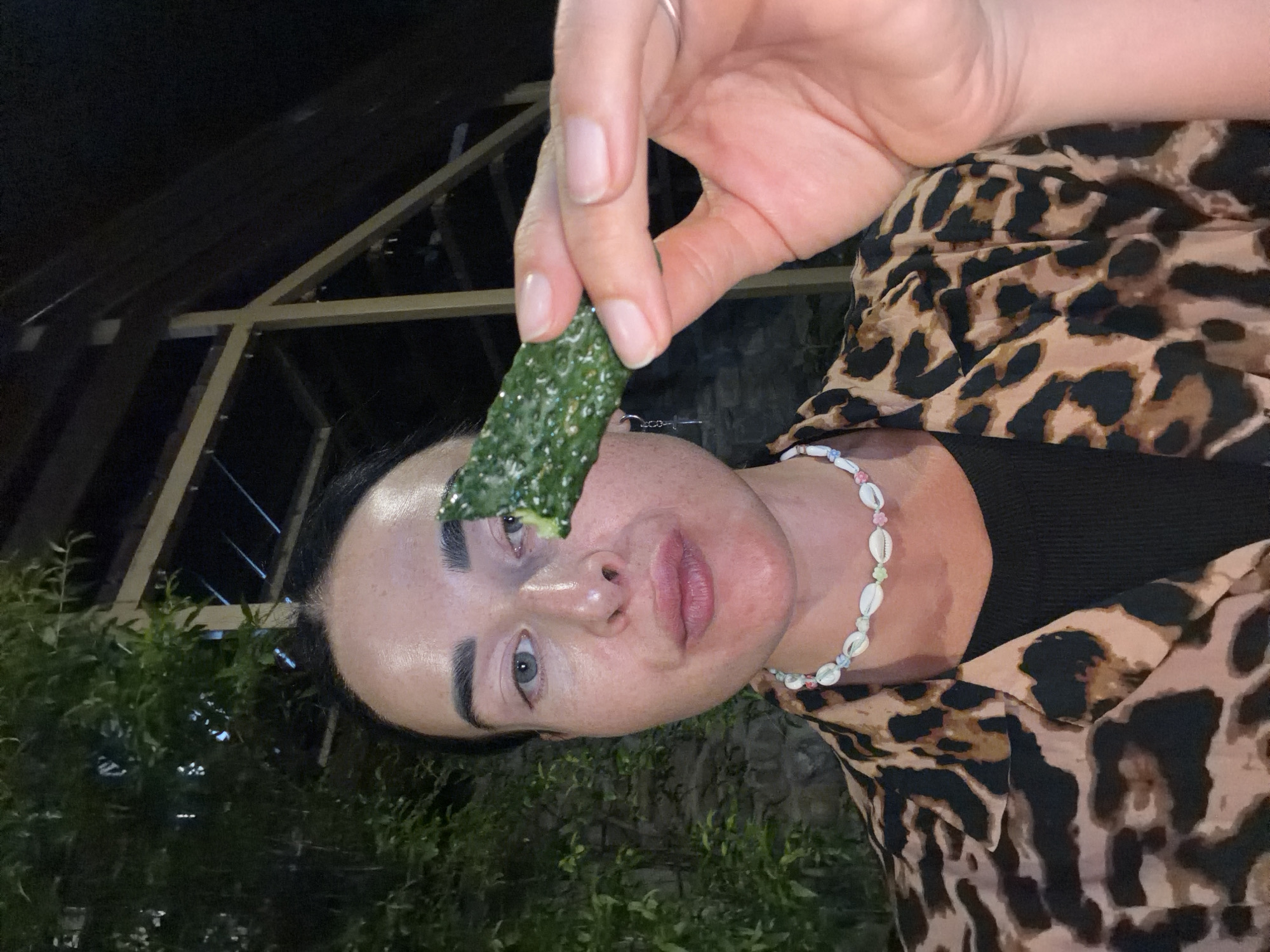 A woman is taking a selfie, holding a piece of cucumber up to her face. She is wearing a leopard print shirt over a black top and a shell necklace. The background shows lush greenery and wooden structures, suggesting an outdoor setting, possibly a restaurant or cafe in Sochi, Russia. The lighting indicates it is nighttime or dusk.