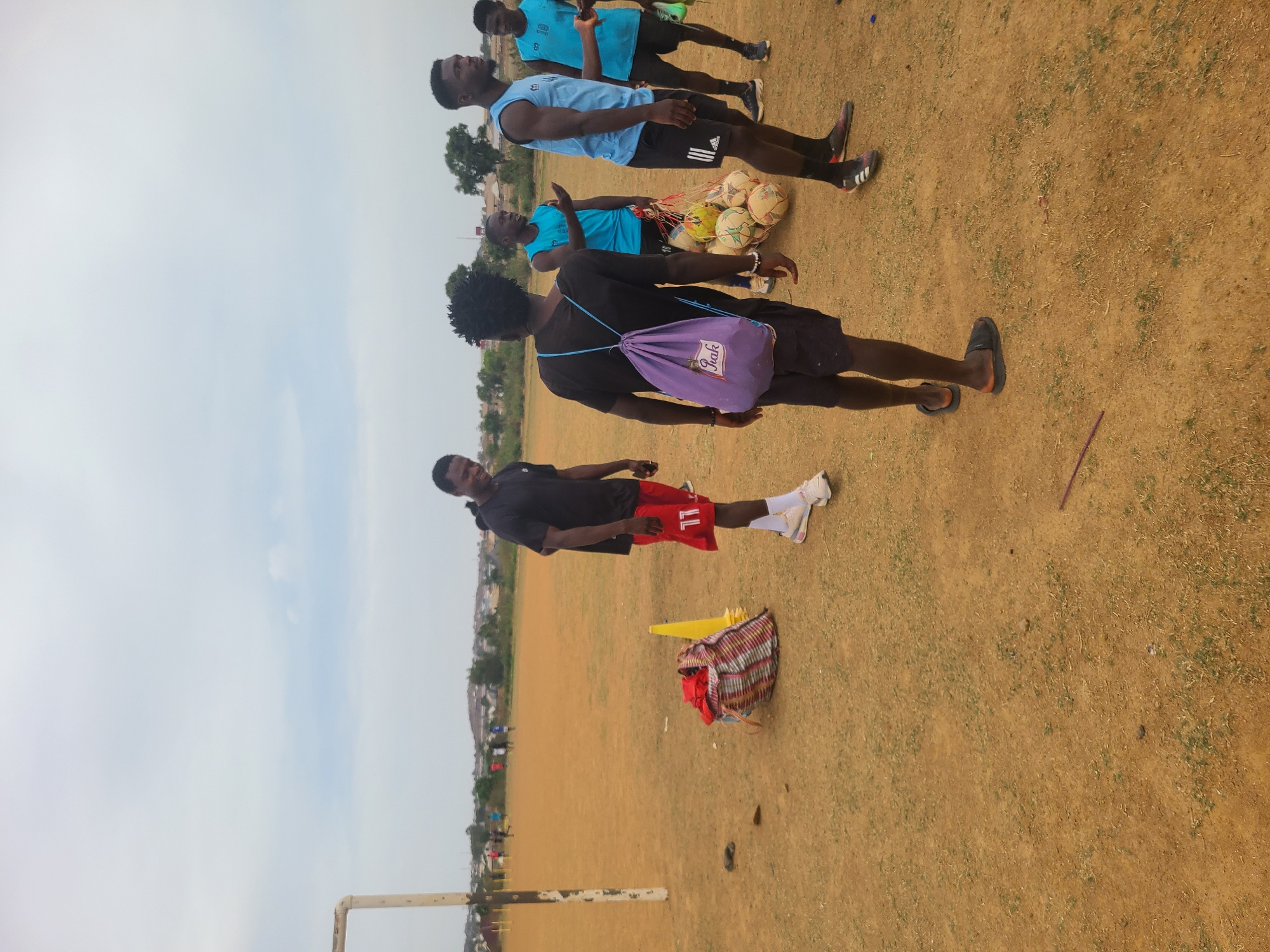 The image depicts an outdoor scene at a soccer field in Barikin Jirga Summa, Nigeria. Several young men are present. Three are wearing light blue jerseys and black shorts. One man is wearing a black shirt and red shorts with the number "11". Another man wears a black shirt and black shorts and carries a purple bag. One person has a net with multiple soccer balls hanging over their shoulder. The ground is predominantly dirt and sparse grass. In the foreground, a yellow cone and a striped bag rest on the ground. A goal post is visible at the edge of the field. The sky is overcast. Buildings are visible in the background.