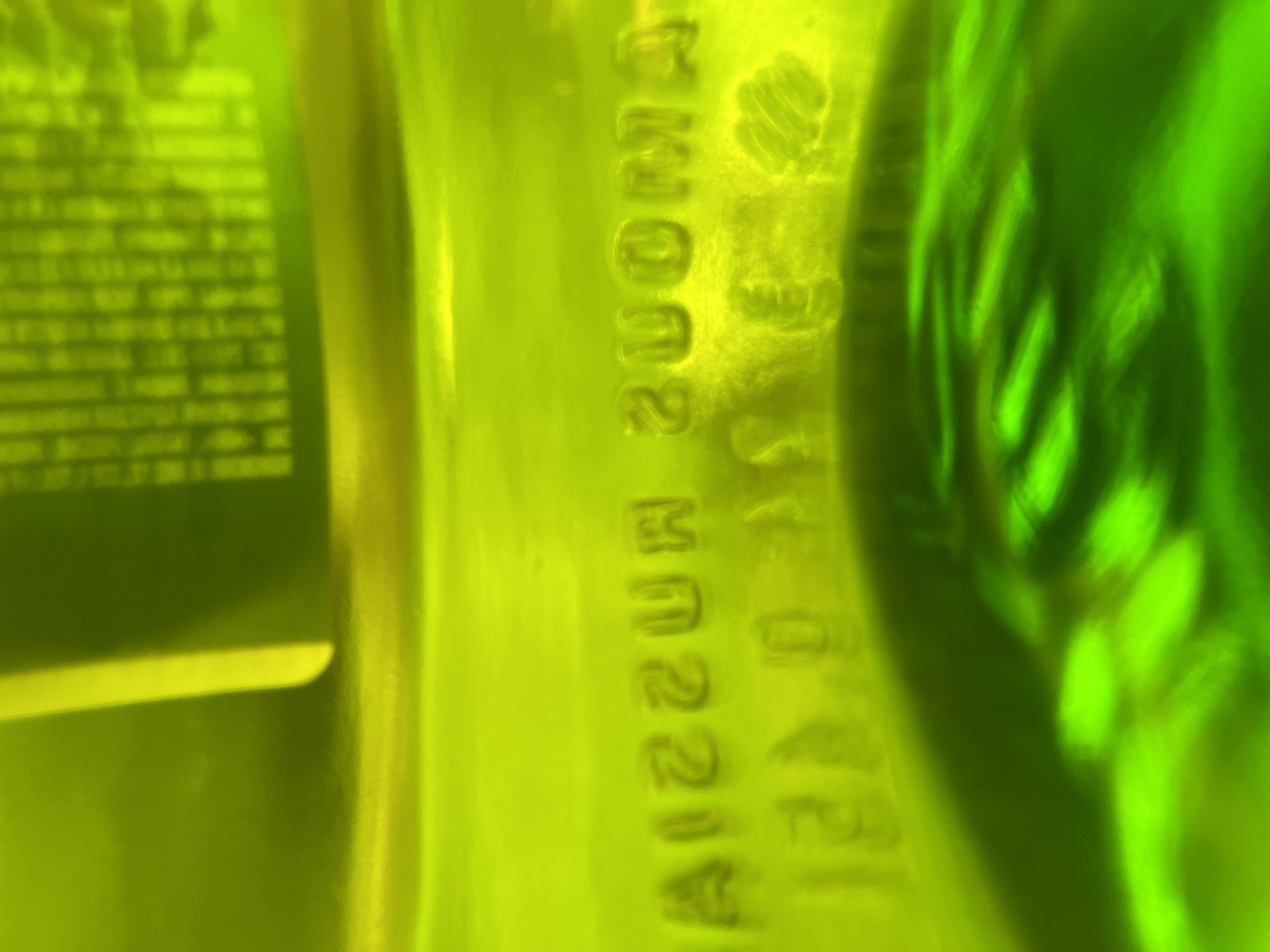 The image displays embossed text on a translucent green object, likely a glass bottle or container. The text appears to be reversed. The upper line of text reads "AIZZUR ZUOMA" and the lower line reads "12.0.4". The object is illuminated from behind, casting a bright green light and creating reflections and distortions. The background is a blurred gradient of yellow and green. There is no visible indication of the city of Vasilyevo, Russia, or any people in the image.
