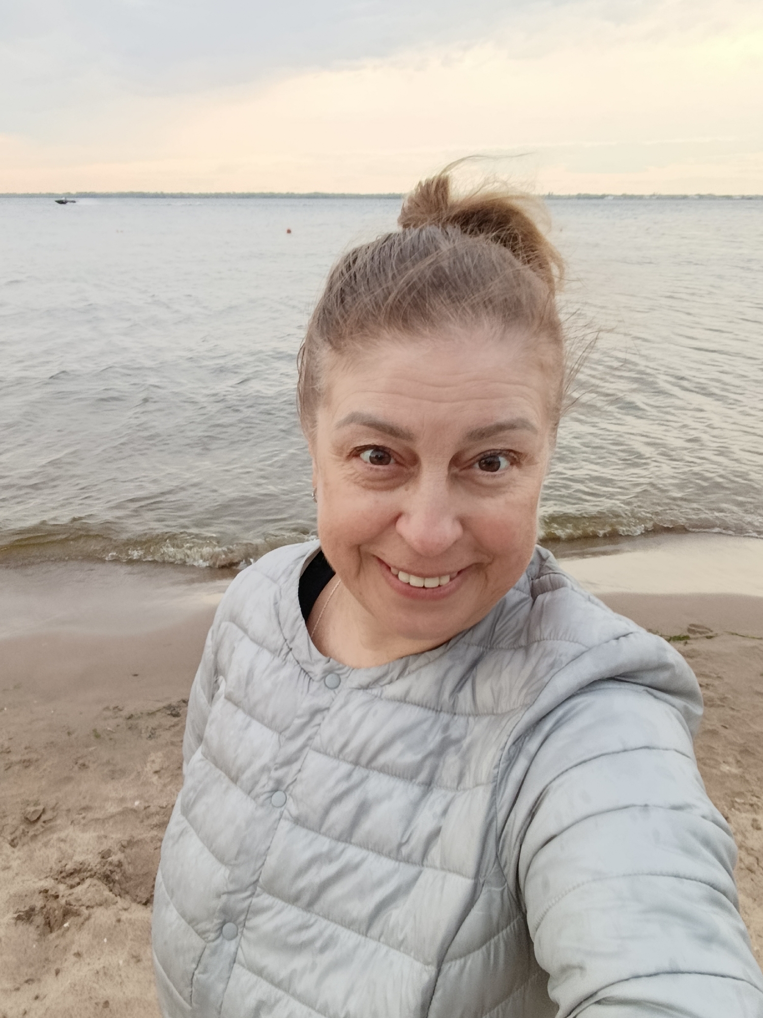 A woman takes a selfie on a beach with the ocean and sky in the background. She is smiling and looking at the camera. She has her hair pulled up in a bun. She is wearing a light gray puffer jacket. The weather appears to be overcast, and the time of day is likely late afternoon or early evening, given the soft light. The beach is sandy, and small waves are lapping at the shore. In the distance, there is a small boat on the water and what appears to be a buoy. The sky is a pale yellow and gray, suggesting a cloudy or twilight setting. The woman's expression is happy and relaxed. There is no visible text in the image. The location context is Saratov, Russia, which suggests the body of water is likely a river or a lake, given Saratov's inland location.
