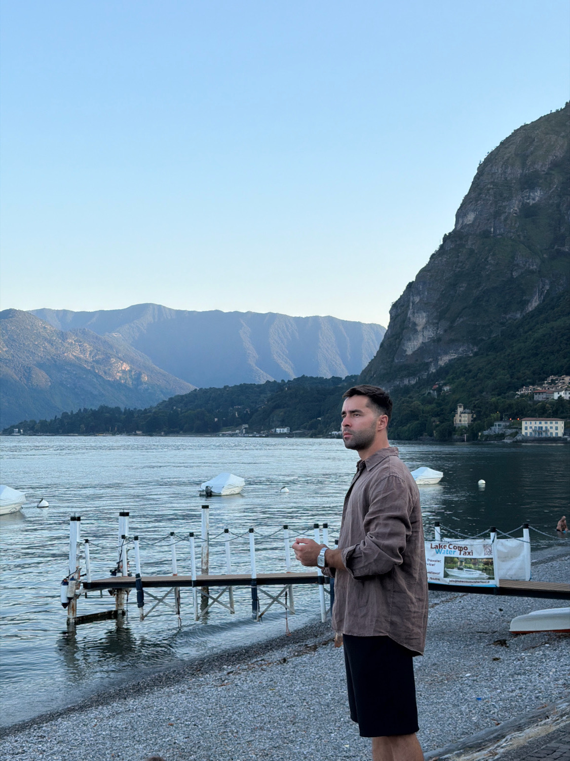 A man stands on a pebble beach in Loveno, Italy, looking out at a calm lake. The man is wearing a brown linen shirt and black shorts. In the background, there are mountains and a few boats on the water. A wooden pier with white railings extends into the lake, and a sign for "Lake Como Water Taxi" is visible. The sky is clear and blue.