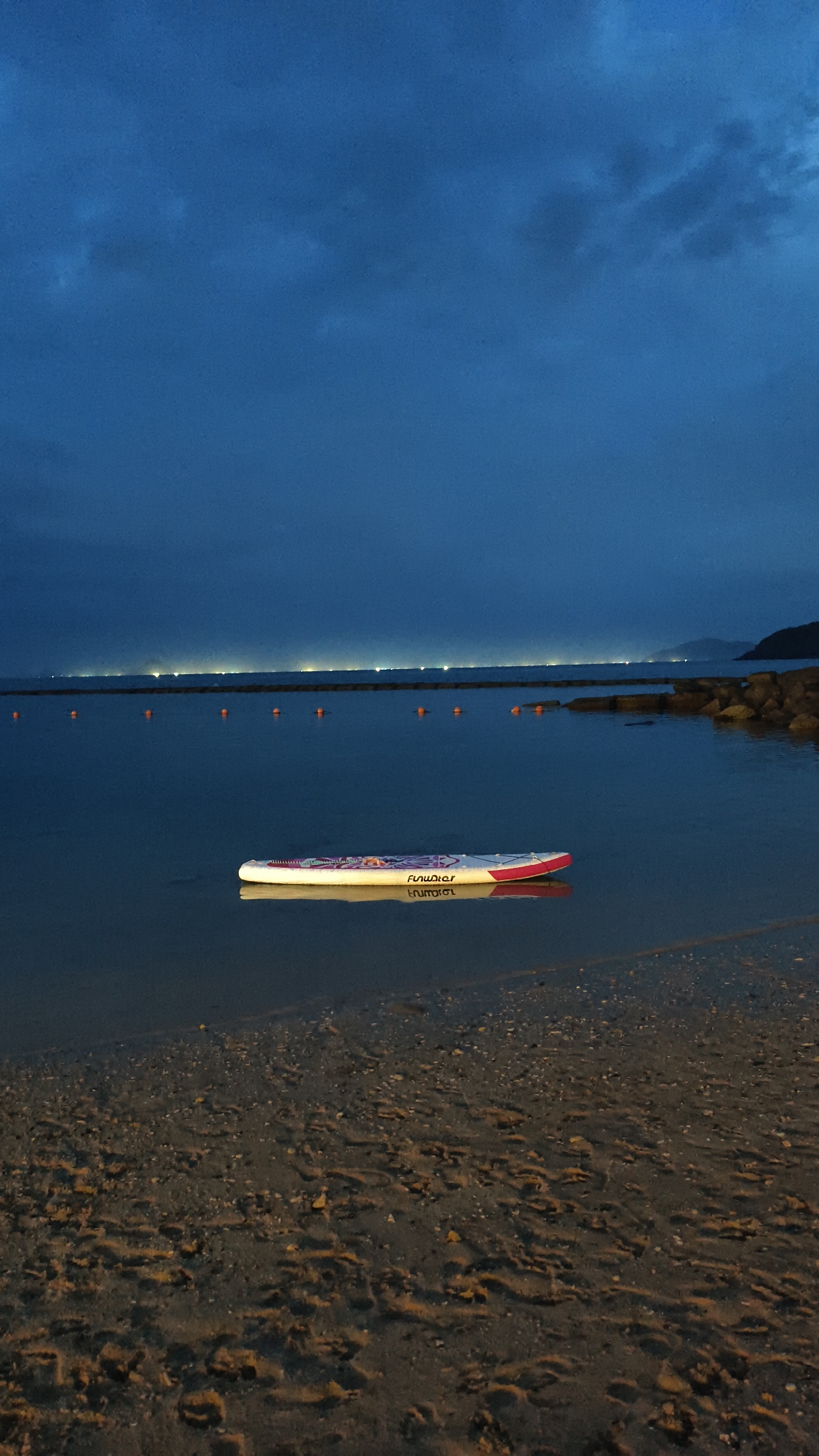 A stand-up paddleboard floats on calm water near a beach at dusk. The water is dark and reflects the dim light from a distant city skyline, which features numerous illuminated buildings. A line of orange buoys stretches across the water, possibly marking a swimming area or a boundary. Rocky structures are visible on the right side of the frame, extending into the water. The foreground shows a sandy beach with textured patterns formed by waves or footprints. The sky is a deep blue, with scattered clouds that are darker and more defined towards the top of the image. The paddleboard is white with red and purple accents and appears to be unoccupied. The overall scene suggests a quiet evening by the sea, likely in a coastal area like Nha Trang, Vietnam, given the presence of distant city lights and a tropical beach setting.