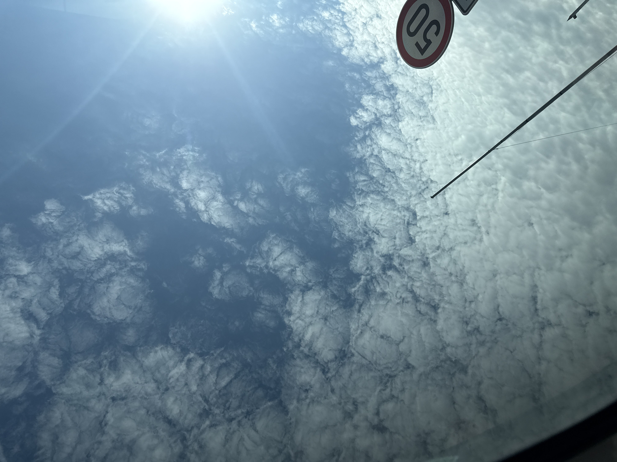 The image captures a bright, sunny day with a blue sky filled with scattered, wispy clouds. In the lower right corner, a circular traffic sign indicating a speed limit of 50 is visible. A slender, dark pole extends diagonally from the lower center towards the upper right. The sun is shining intensely from the upper right, creating a lens flare. The perspective suggests the photo was taken from inside a vehicle, looking upwards through a windshield, with a sliver of the car's interior framing the lower left edge. The scene does not depict any people or specific city landmarks that would allow for identification of Varna, Bulgaria.