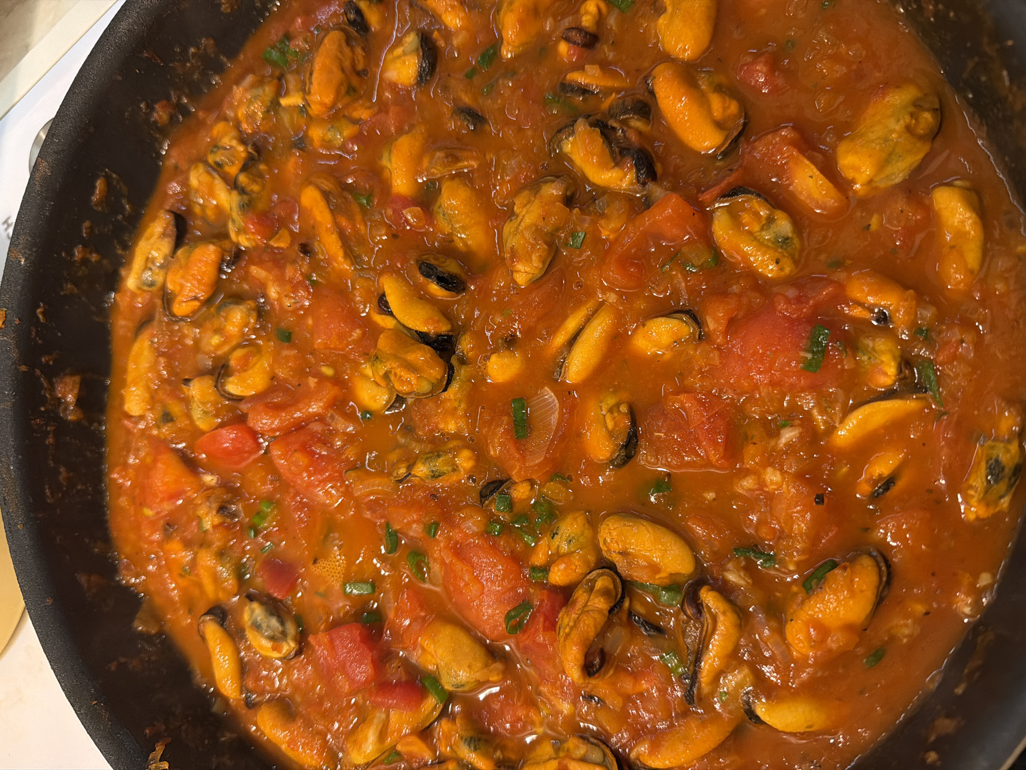 This overhead, close-up shot features a black cooking pan filled with a vibrant seafood dish. The main subjects are numerous shelled mussels, exhibiting a bright orange-yellow hue, immersed in a rich, reddish-orange tomato-based sauce.
The sauce is thick and chunky, containing visibly substantial pieces of red tomato and translucent slivers of what appear to be onions. Small fragments of chopped green herbs, possibly parsley or chives, are sprinkled throughout, adding a fresh contrast to the warm colors of the sauce and mussels.
The scene captures the food either actively simmering or having just been prepared, indicated by the glistening surface of the sauce. The cooking vessel is a dark, possibly non-stick, pan. A small portion of a white surface, likely a stovetop or counter, is visible along the left and bottom-left edges of the frame.
There are no people present, and no specific event beyond the preparation or serving of food. The image provides no direct cues for time of day or weather. No text is visible within the frame. The overall impression is one of a robust and flavorful meal, focusing solely on the culinary elements.