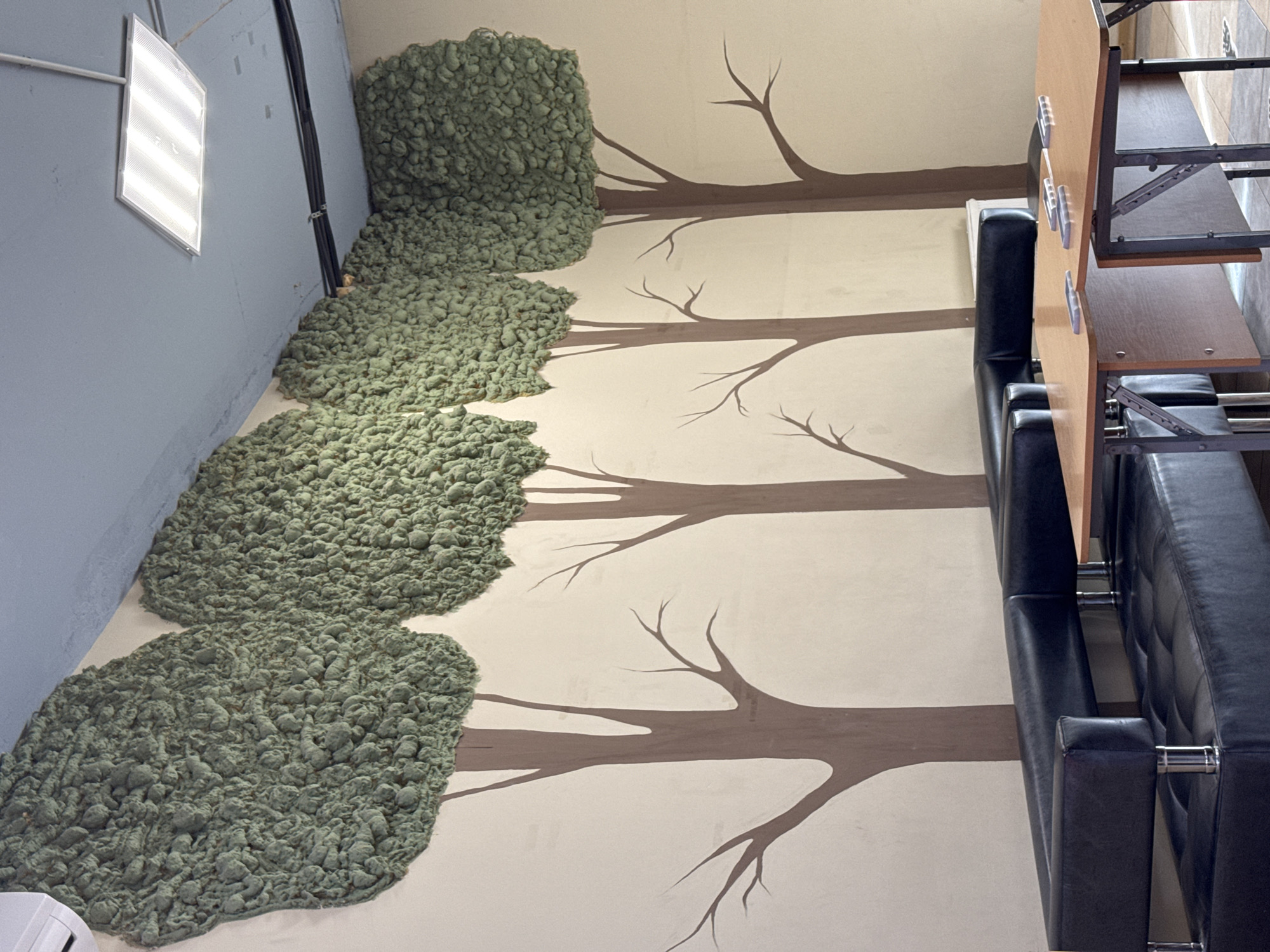 The image shows an interior scene, likely an office or meeting room, with a distinctive wall decoration resembling trees made of green textured material. The wall itself is a light, neutral color. In the foreground, a long, black leather sectional sofa is visible. In front of the sofa, two wooden tables are placed side-by-side. The tables appear to be set up for a meeting, with several transparent plastic objects, possibly card holders or placemats, arranged on their surfaces. The ceiling is painted blue and features a fluorescent light fixture. The environment appears to be a modern indoor space. No people are present in the image. The city of Perm, Russia is mentioned as the location, but there are no specific visual cues within the image to confirm this.