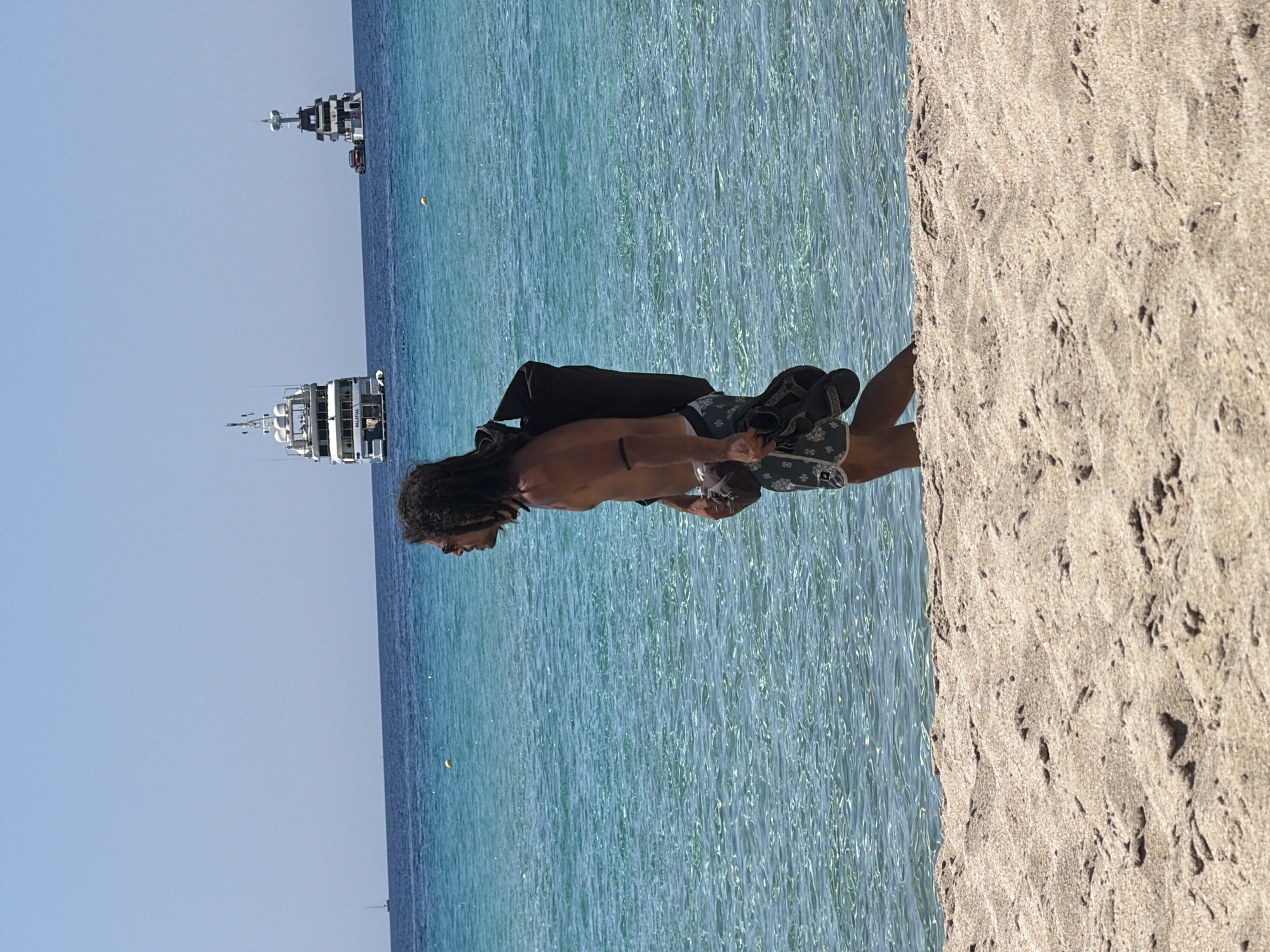 A man with long, dark, wavy hair and sunglasses is walking along the beach in Greece. He is shirtless and wearing patterned shorts. He is carrying a black backpack over his shoulder and a pair of dark sandals in his right hand. The ocean water is a clear turquoise color, and several large yachts are visible in the distance. The beach is sandy, with the foreground dominated by sand dunes. The sky is clear and blue.