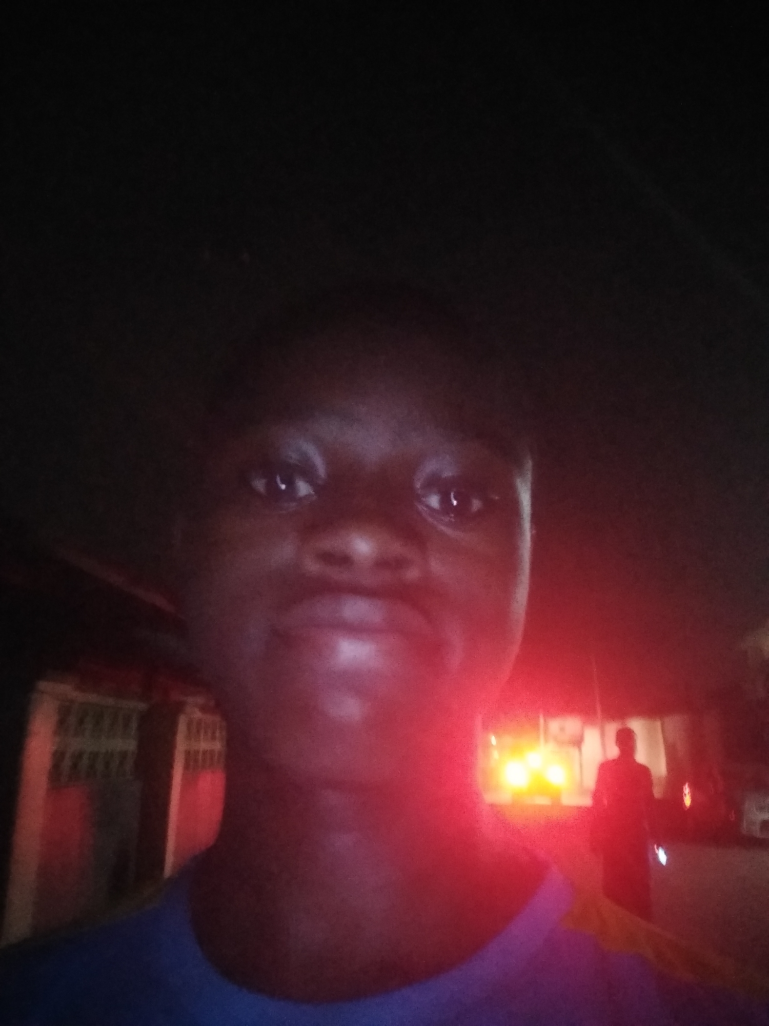 This is a night-time selfie taken in Shangisha, Nigeria. The primary subject is a young person, positioned centrally and dominating the foreground, looking directly at the camera. Their face and upper body are illuminated by a strong red light emanating from the right side of the frame, casting a reddish glow on their skin and a blue garment (likely a t-shirt or shirt) they are wearing. Their expression appears neutral, with slightly pursed lips.
In the blurred background, to the right, a silhouetted figure of another person is visible, walking or standing. Behind this figure, several bright red and yellow lights are prominent, suggesting the presence of vehicles or emergency lights. On the left side of the background, a section of a wall or building with a horizontal patterned structure (possibly concrete blocks or a fence) is partially visible, also caught in the red illumination. The overall darkness of the image confirms it was taken at night. No visible text is present.