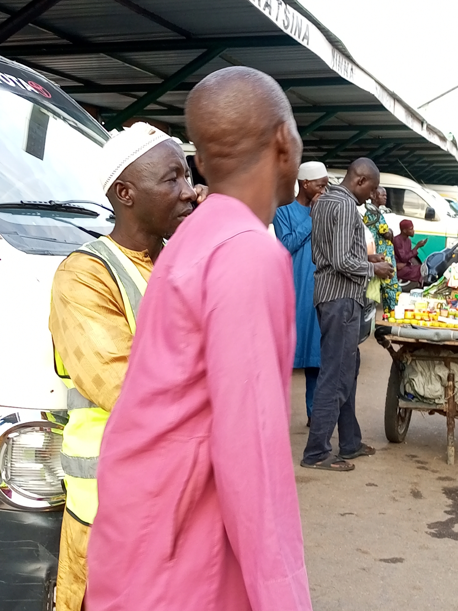 The image depicts an outdoor scene, possibly a market or a transportation hub, in Gombe, Nigeria. In the foreground, a person wearing a bright pink garment is partially visible. To their left, an individual in a gold-colored robe and a high-visibility vest is standing next to a white vehicle. Further back, several other individuals are present. One man wears a blue outfit, and another is in a striped shirt and dark trousers. A woman in a colorful patterned dress is also visible, as is a person in a dark red garment. In the background, a covered structure with a corrugated roof is visible, and a cart laden with various items, including what appear to be jars and bottles, stands on the ground. The ground surface appears to be paved, with some wet patches.