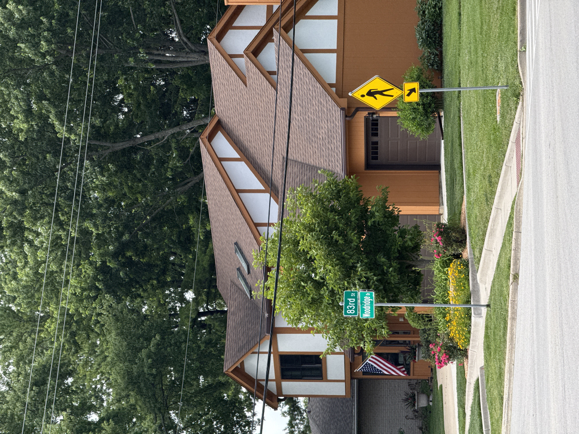 The image shows a residential street in Woodridge, United States of America. In the foreground, a paved road curves to the left, bordered by manicured green lawns and concrete curbs. Two street signs are visible: one green sign reads "83rd St" and "Woodridge Dr," indicating the intersection of these two streets. Above it, a yellow diamond-shaped sign depicts a pedestrian silhouette, a common "pedestrian crossing" warning sign. Below that, another yellow sign shows a black arrow pointing to the right.
A two-story house with a brown roof and distinctive orange siding dominates the midground. The house features white stucco panels with brown trim, and a dark brown garage door is visible on the right side. An American flag is displayed on the left side of the house. A mature tree with lush green foliage stands prominently in front of the house, partially obscuring its facade. Flower beds with pink and yellow blossoms are planted along the curb in front of the house.
In the background, a dense canopy of green trees forms a natural backdrop against a cloudy sky. Power lines are strung across the upper portion of the image, traversing in front of the trees. The overall scene depicts a well-maintained suburban environment.