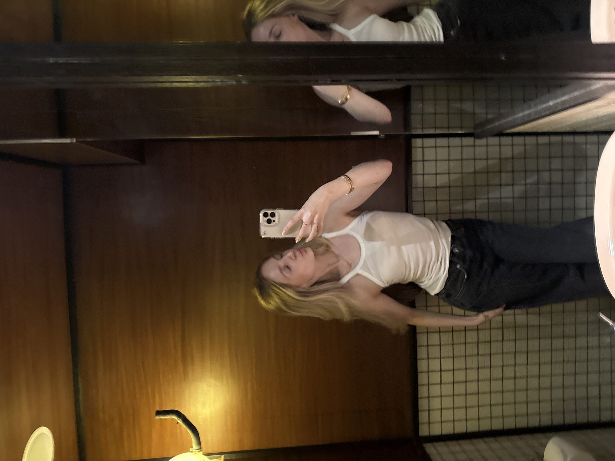 A young woman with long blonde hair is depicted taking a mirror selfie indoors. She is reflected in a large mirror, holding a light-colored smartphone with her right hand to capture the image. She wears a white ribbed tank top and dark jeans. A ring is visible on her right hand, and she has a relaxed expression with her lips slightly parted.
The setting appears to be a room with dark wooden paneling or walls, suggesting warm and somewhat dim lighting. Above the woman's reflection, a dark shelf or beam is visible, along with another reflection of a woman's face, possibly from a different mirror or angle. To her right, a black grid-like pattern is visible on a wall or surface, alongside part of a white sink or basin. A dim light fixture with a curved black pipe is partially visible in the lower left of the reflected scene. The orientation of the original image shows the woman lying on her side. No discernible text is visible in the image.
