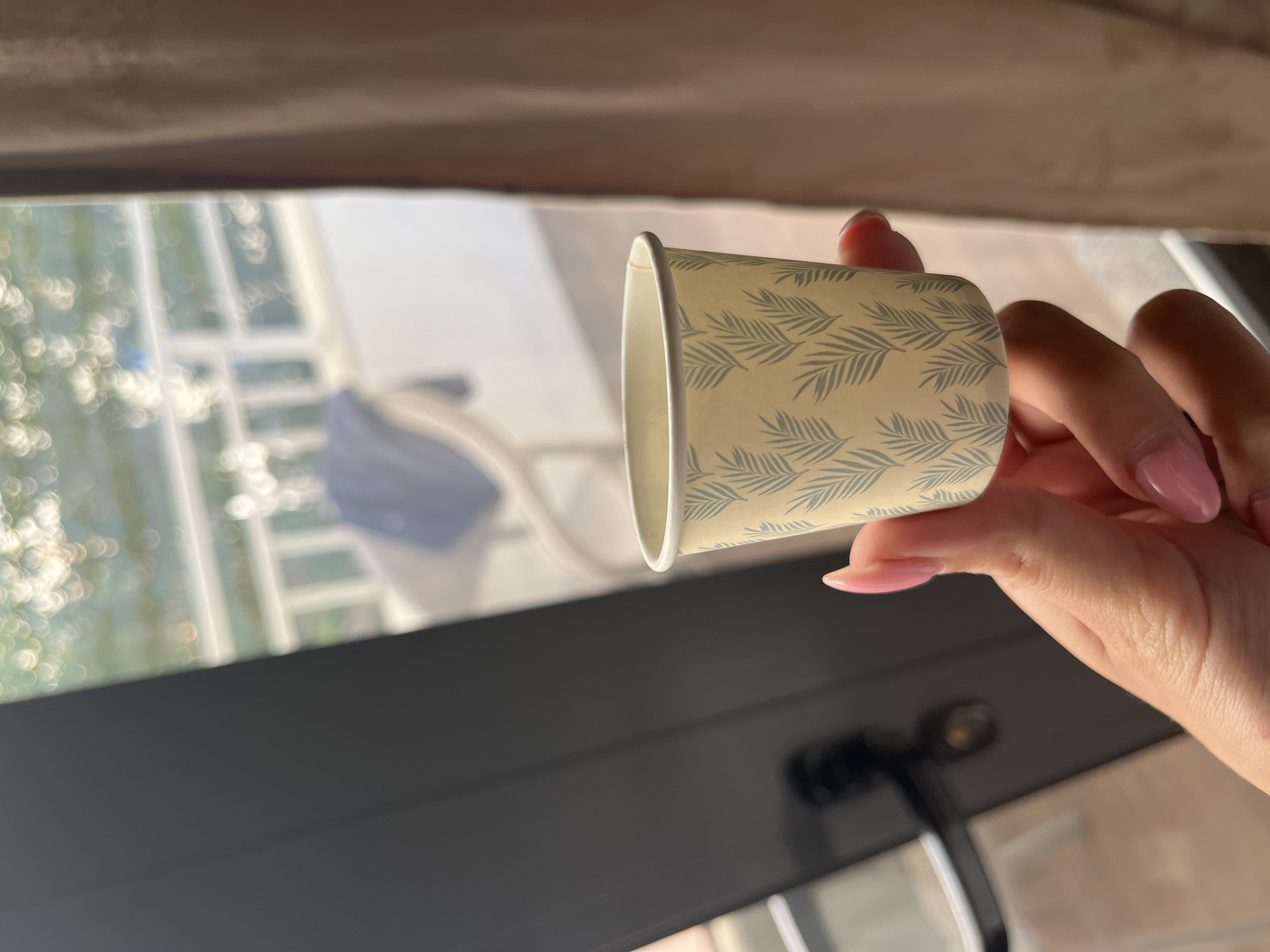 A close-up shot shows a person's hand holding a small, patterned paper cup. The cup has a pale yellow background with a repeating pattern of gray palm leaves. The hand has manicured, light pink nails and is positioned in the foreground.
In the background, out of focus, is a balcony with a white railing, suggesting an outdoor setting. A chair with a dark blue cushion is visible on the balcony. The overall lighting is soft, with bokeh effects from light sources reflecting on the balcony's surface. A dark vertical object, likely a door or window frame, occupies the left side of the image. A textured, light brown curtain is visible on the right.
The specific location in Bashkia Durrës, Albania cannot be determined from the visual information alone.