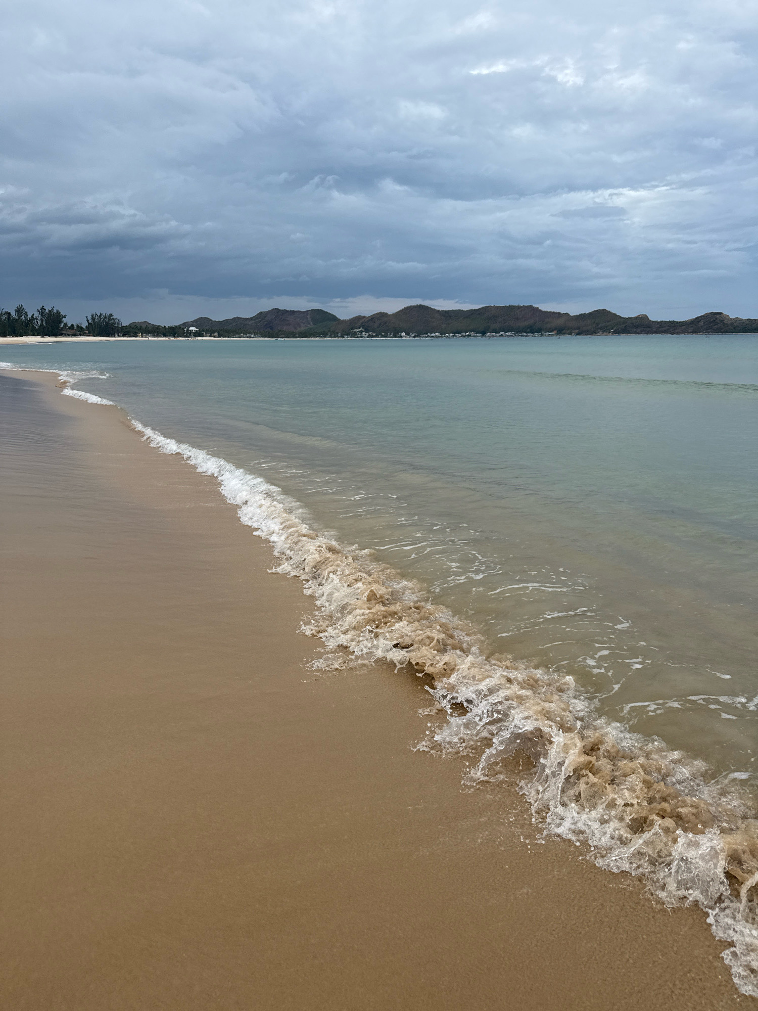 The image shows a serene beach scene in Song Cau, Vietnam, under a cloudy sky. The foreground features a sandy beach with gentle waves lapping at the shore. The water is a light, clear blue, contrasting with the darker, muted tones of the distant hills and the overcast sky. A line of trees is visible along the coastline, with some structures nestled among them, suggesting a settlement or resort area. The overall atmosphere is calm and peaceful, with no people visible in the frame.