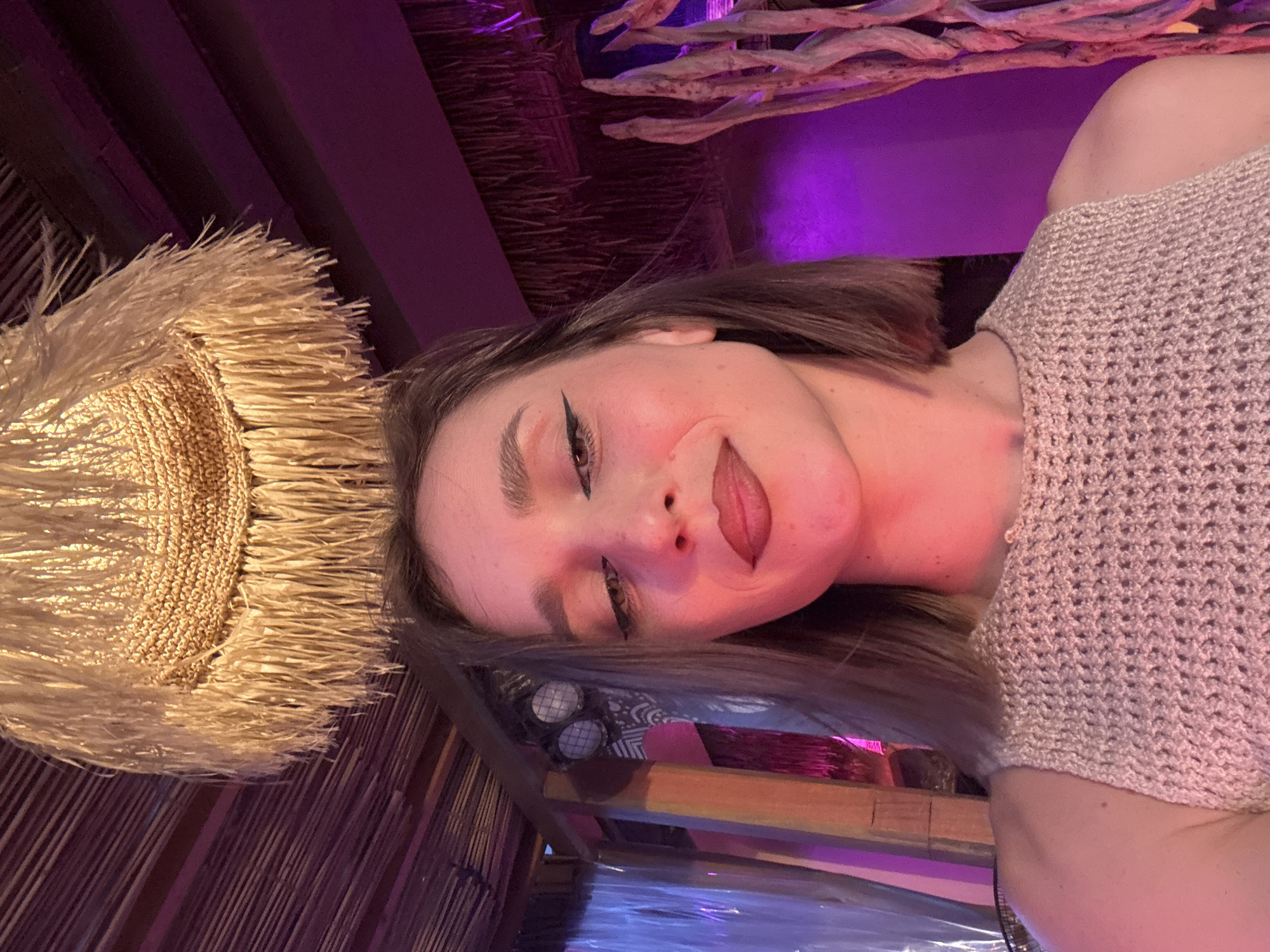 In an indoor scene with purple lighting, a young woman with brown hair is captured in a selfie. She is wearing a beige crocheted top. Above her, a straw lampshade with fringe hangs from a wooden ceiling with a diagonal striped pattern. The background features pink and purple lighting, along with wooden structural elements and what appear to be dried branches.