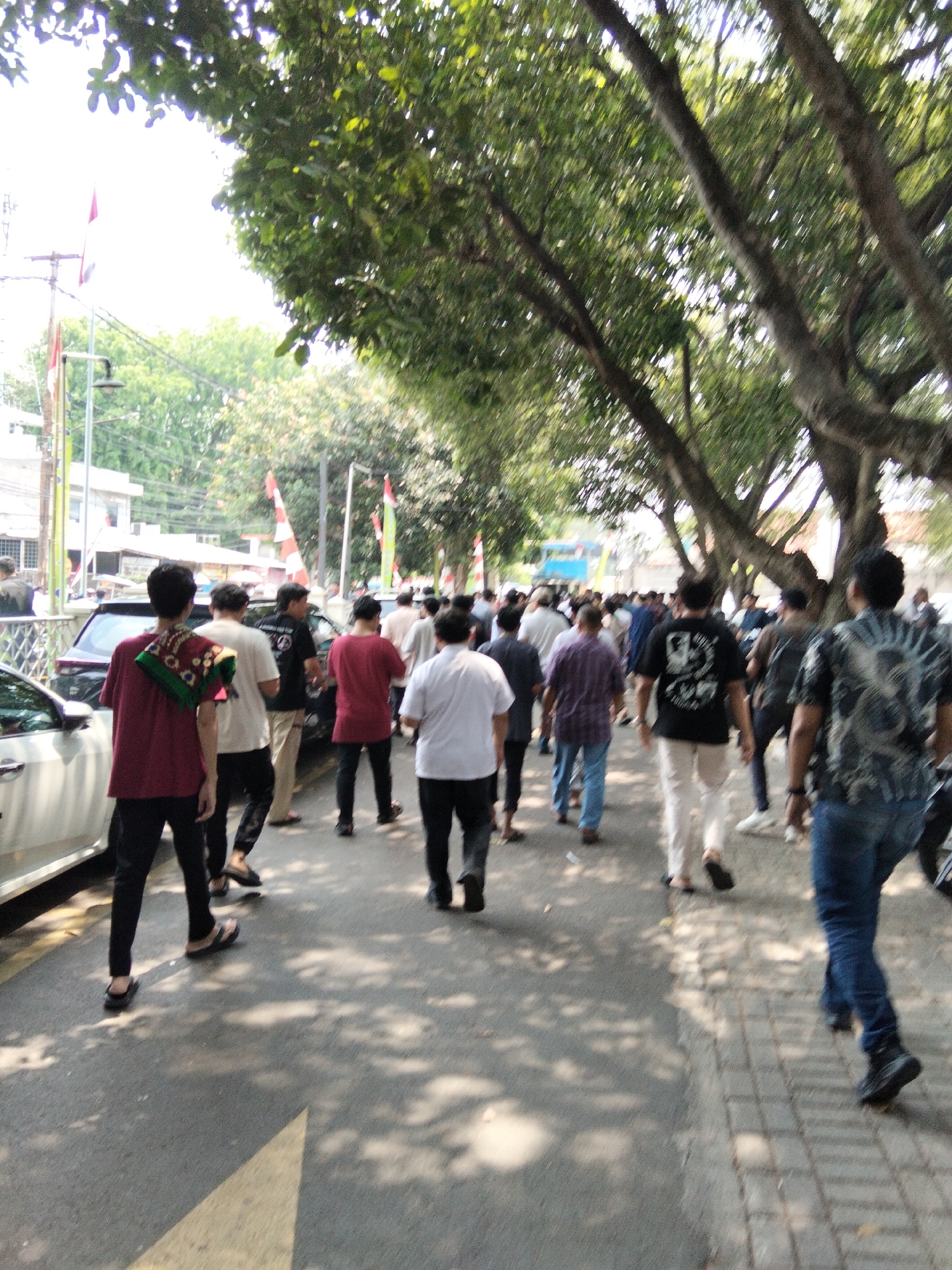 The image depicts a group of people walking along a tree-lined street in Depok, Indonesia. The scene appears to be outdoors, with sunlight filtering through the leaves of large trees casting shadows on the pavement. Several individuals are visible, walking in the same direction. Some are dressed in casual clothing, while others wear more formal attire. Flags, possibly national or organizational flags, are visible in the background, suggesting a public event or gathering. Cars are parked along the side of the road, and buildings can be seen in the distance. The overall atmosphere suggests a bustling urban environment.