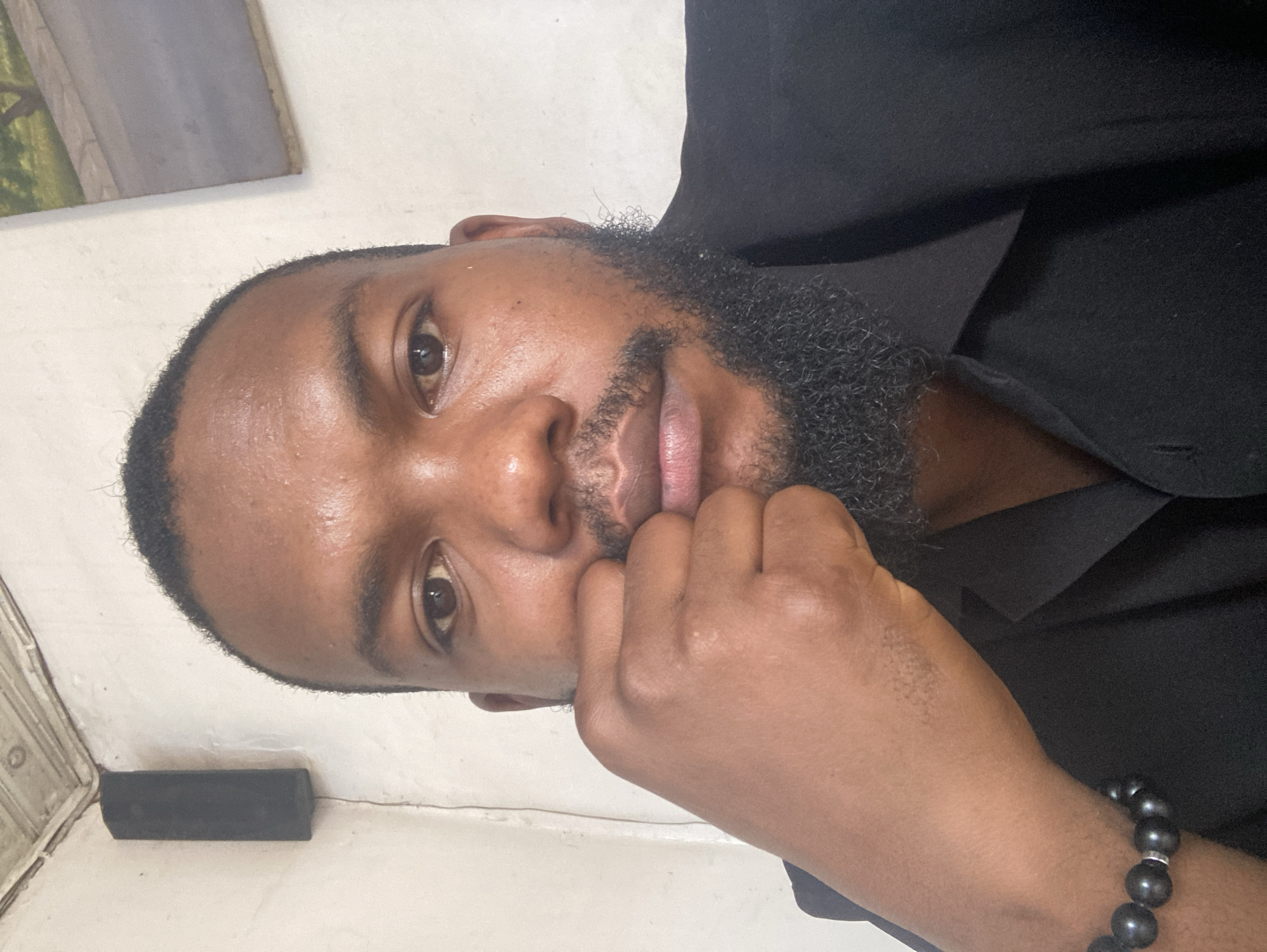A close-up, slightly rotated portrait shows a man with dark skin, a beard, and a short haircut, his face mostly visible and his right hand positioned near his chin in a contemplative pose. He is wearing a black shirt, and a beaded bracelet is visible on his wrist. The background is plain, possibly a wall with a framed picture partially visible in the upper left corner. There is no information in the image that allows for identification of Gwong, Nigeria as the specific location.