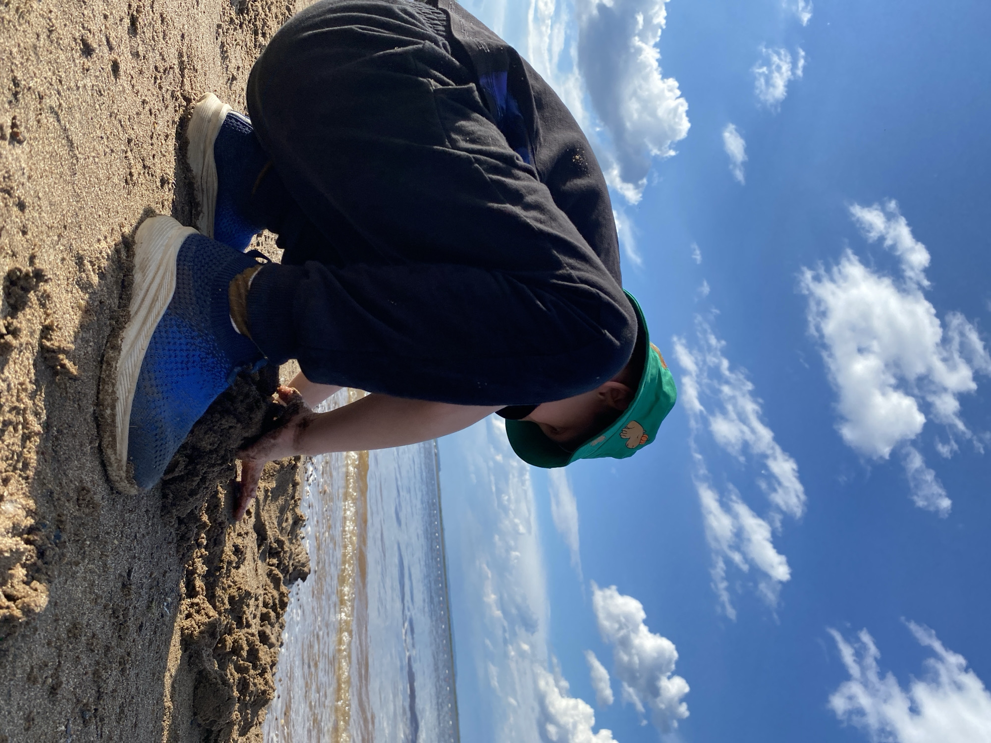 A child wearing a green bucket hat with a capybara design and blue sneakers is playing in the sand near the water. The child is crouched down, digging in the sand with their hands. The sky is bright blue with fluffy white clouds. The water is calm with small waves gently lapping at the shore. The scene evokes a sense of childhood fun and summer enjoyment.