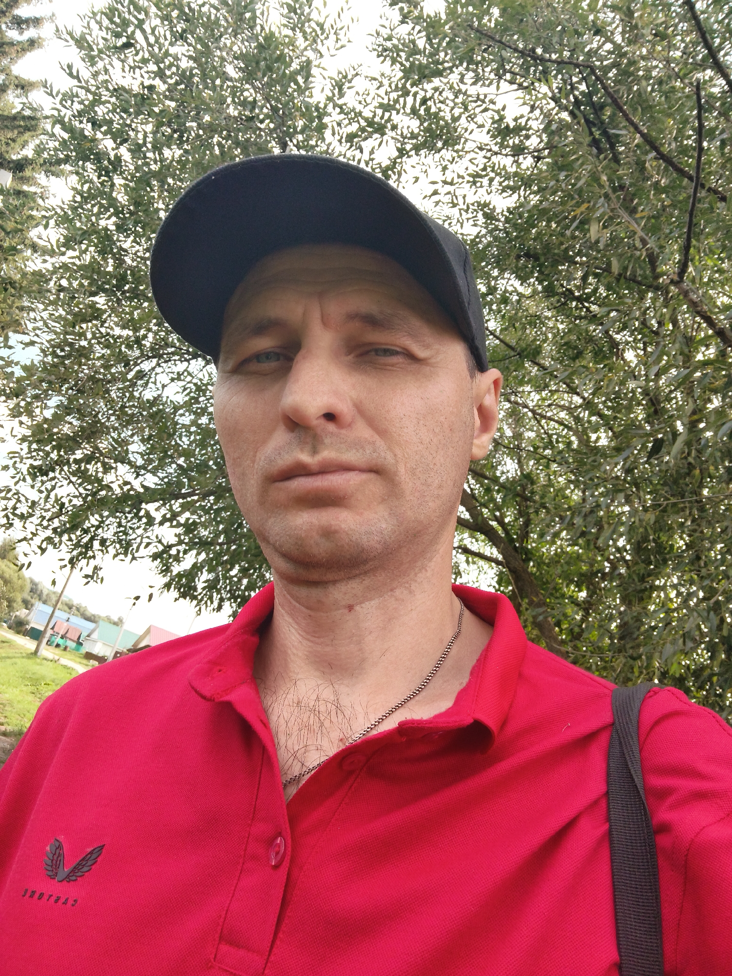 A middle-aged Caucasian man wearing a red polo shirt and a black baseball cap is pictured from the chest up, facing the camera. He has short, light brown hair and a fair complexion. He is wearing a silver chain around his neck. In the background, there are trees with green leaves, and a hint of buildings and a grassy area in the distance, suggesting an outdoor, possibly rural or suburban setting. The image quality is decent, but there is some graininess. The lighting suggests it is daytime. The man is not actively engaged in any discernible action, and the setting does not provide specific clues to a precise location within Mokshan, Russia, beyond a general outdoor environment.