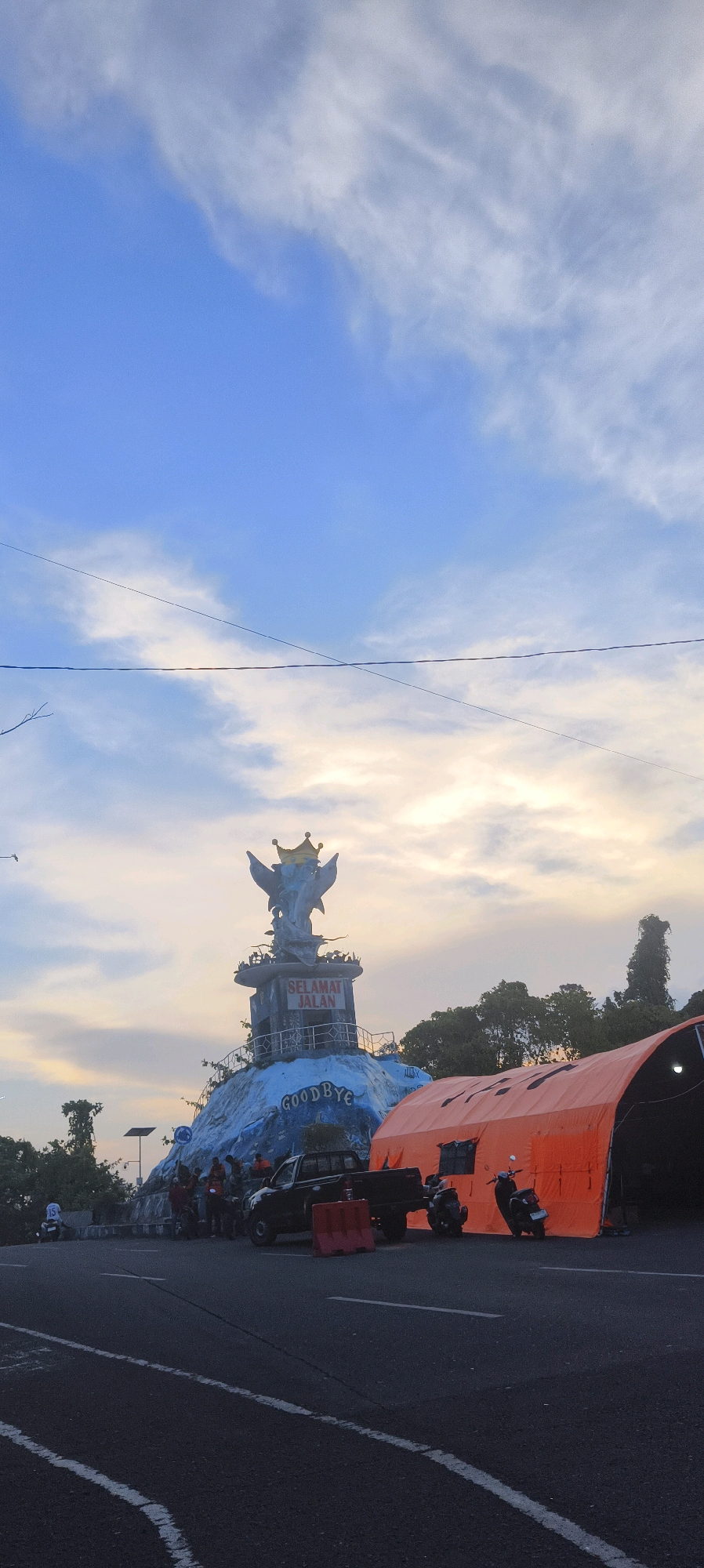 The image captures a monument and an orange tent structure in the city of Waisai, Indonesia. The monument features a sculpture of dolphins crowned with a golden headdress, positioned on a tiered platform. The platform displays text that reads "SELAMAT JALAN" on a sign and "GOODBYE" on the lower portion. The monument is situated on a mound with a blue, rocky-textured base.
In the foreground, a road is visible with white lane markings. Adjacent to the road, several individuals are gathered near a black pickup truck and motorcycles. To the right of the monument, a large orange tent is erected, appearing to be a temporary structure. The sky above is a mix of blue with scattered white clouds, suggesting it might be late afternoon or early morning, with warm light filtering through. Trees are visible in the background, some appearing tall and slender, others more bushy. Power lines are also present, stretching across the sky.