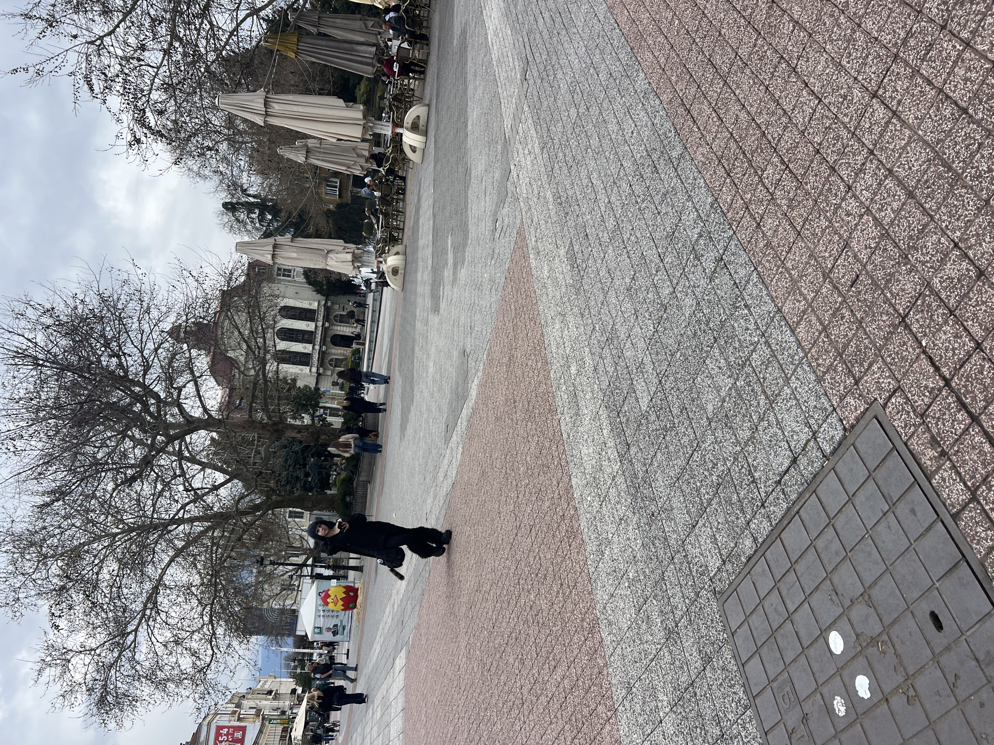 The image depicts a public square in Plovdiv, Bulgaria. The square is paved with gray and red interlocking paving stones. A woman dressed in black is walking diagonally across the square towards the right side of the frame. She appears to be holding a mobile phone. A large, bare tree is to the left of the woman. In the background, there is a building with white columns and a red roof. There are several people seated at tables with open umbrellas. A utility access cover is on the right side of the frame.
