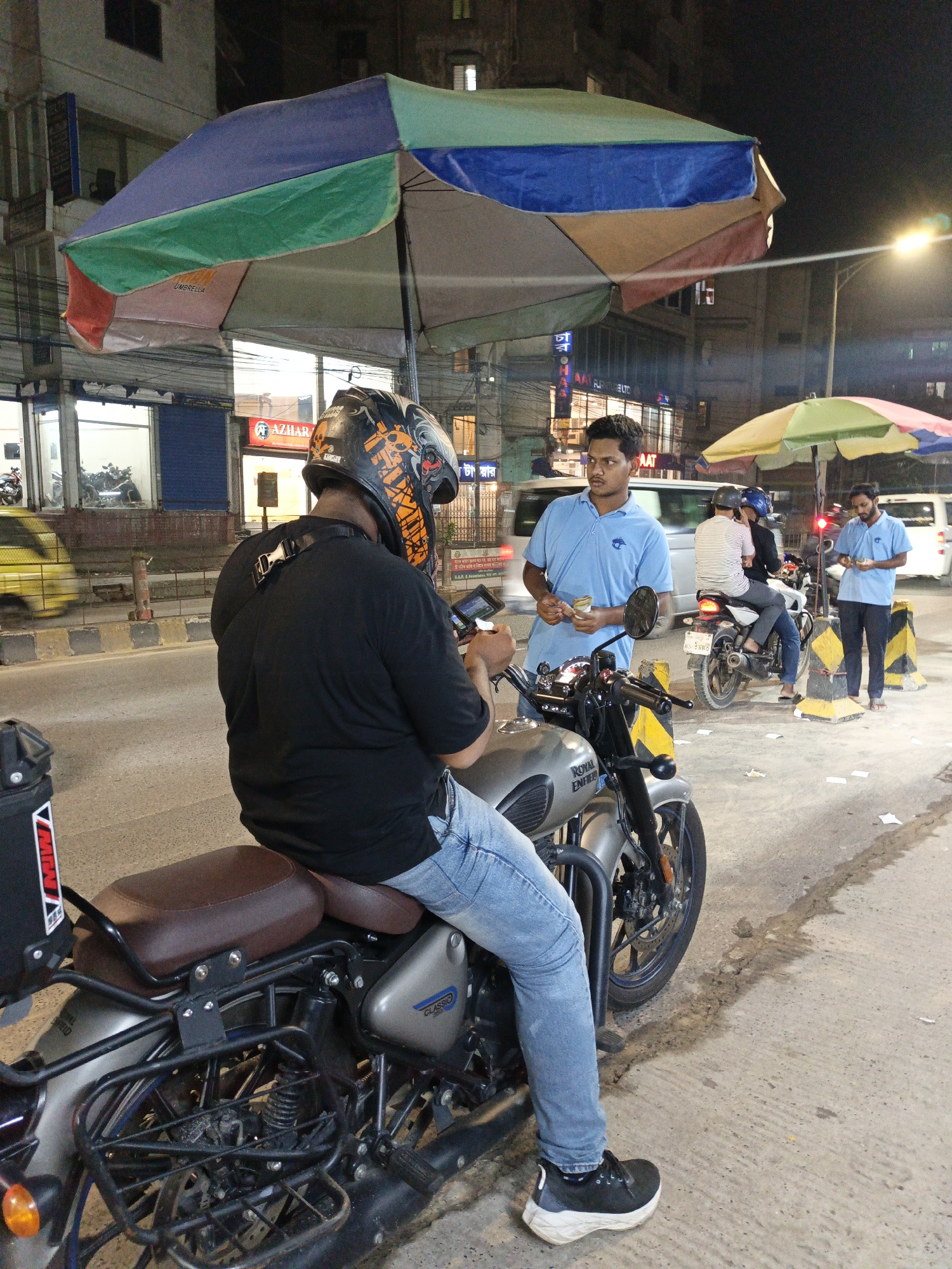 At night in an urban environment identified as Unknown, Unknown, a person wearing a black t-shirt, light blue jeans, and black and white sneakers is seated on a gray Royal Enfield motorcycle under a large, multicolored umbrella. The person is wearing a black helmet with orange and white graphics and appears to be looking at a mobile phone. A second man, wearing a light blue polo shirt, stands next to the motorcycle holding money in his hands. In the background, there is another pair of individuals on a motorcycle, also associated with men in light blue polo shirts, under another smaller umbrella. There are multiple vehicles and buildings visible in the background, including shops with signage, and streetlights illuminate the scene. The ground shows signs of wear and tear, including dark streaks and scattered debris.