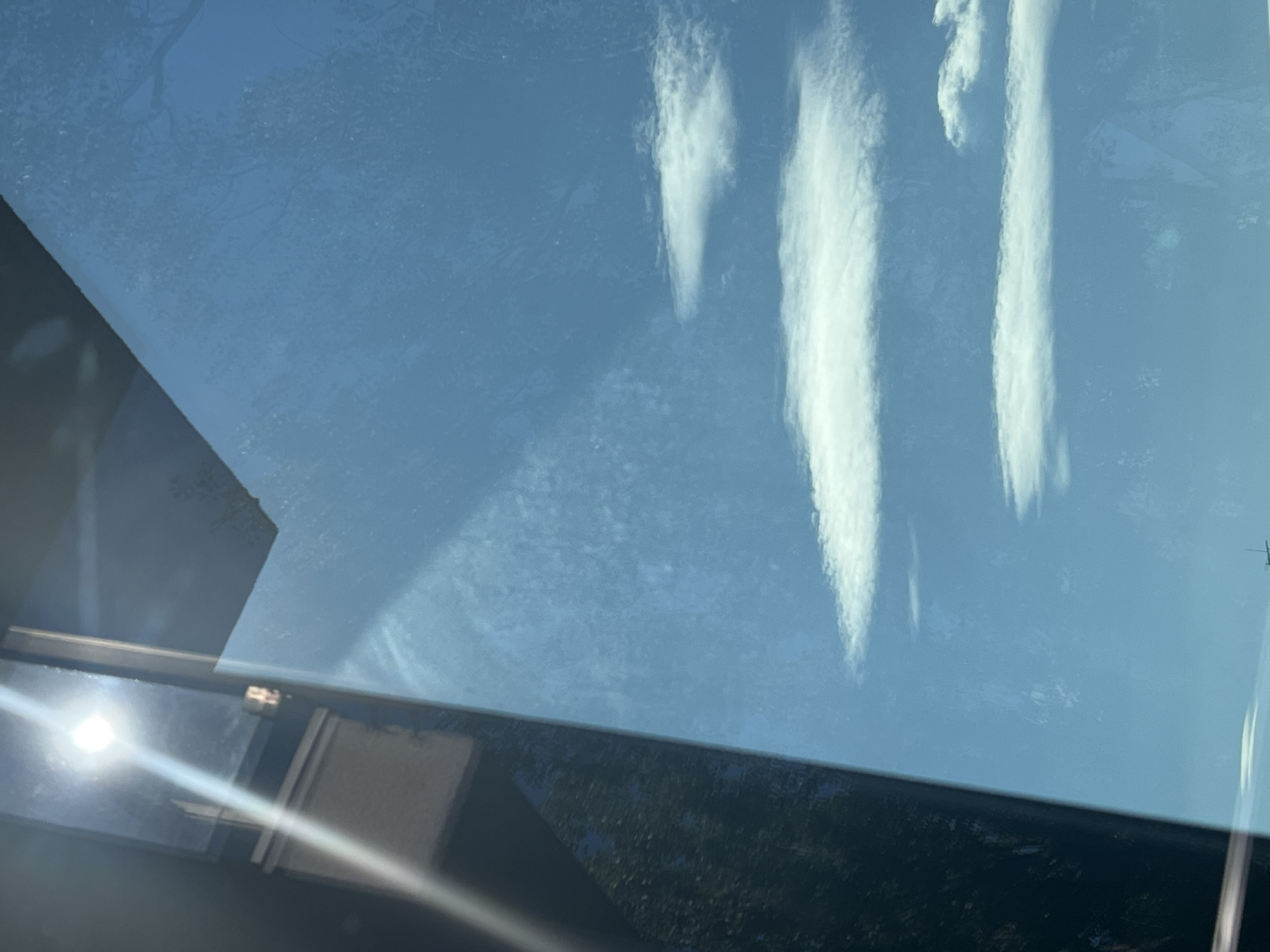 The image shows a bright sun shining through what appears to be a window, with a reflection on the glass of trees and sky. The sky is blue and there are a few white clouds scattered across it. The sun is a bright white orb with rays of light emanating from it. There is a dark, vertical object on the left side of the image that could be a window frame or part of a building. The scene is bright and appears to be taken during the daytime. There is no discernible information within the image to suggest a specific location in Warsaw, Poland.