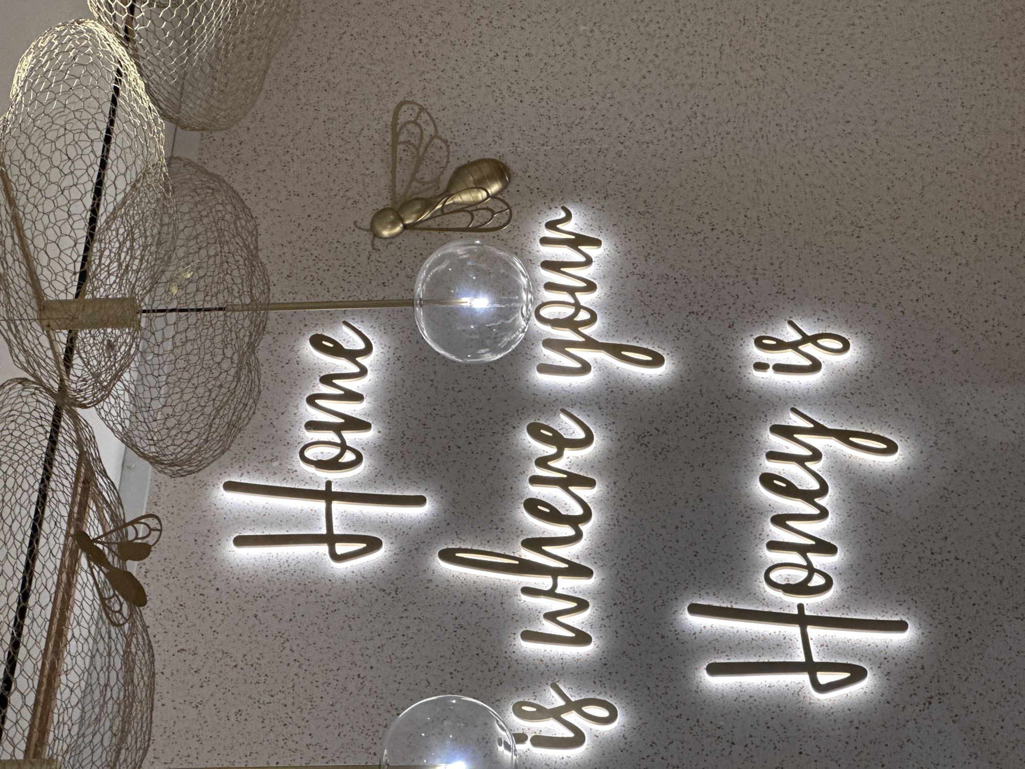 The image shows a ceiling decoration in a room. The ceiling is a light gray speckled pattern. Attached to the ceiling are lit-up, gold-colored script letters spelling out "Home is where your Honey is". There is a gold-colored metal bee and what appear to be glass ball light fixtures. To the left of the phrase is a light fixture with a wire mesh outer layer. The image was taken looking up at the ceiling. The image does not contain any information about the location in London, United Kingdom.