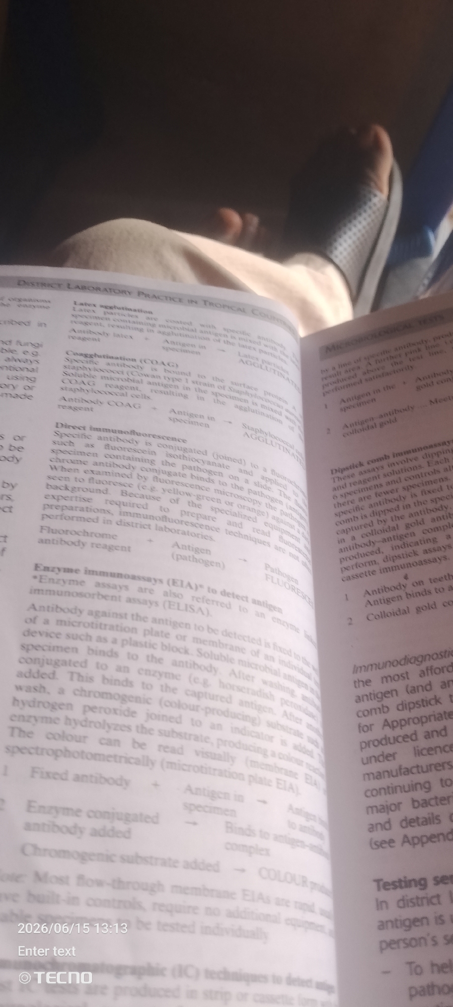 The image shows a close-up, overhead view of an open textbook displaying information on laboratory practices and microbiological tests. The text is the primary subject, with some of the lower right corner of the image showing a person's legs and feet, wearing slippers and resting on what appears to be a soft surface. The environment is dimly lit, suggesting an indoor setting. There are no visible elements that would indicate the location of Jalingo, Nigeria. The scene is static and focuses on the academic material presented in the book.