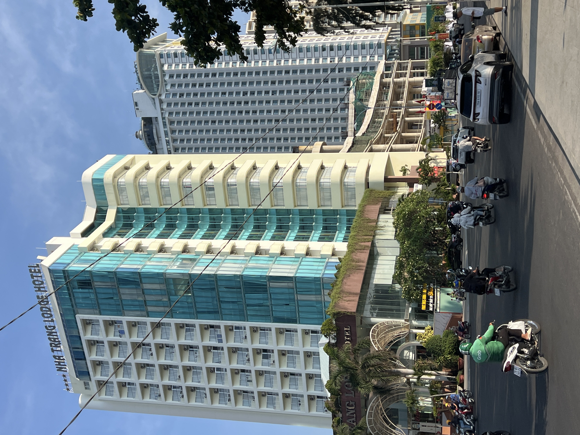 The image depicts a street scene in Nha Trang, Vietnam, prominently featuring the Nha Trang Lodge Hotel. The hotel is a multi-story building with a mix of light yellow and blue glass facade. The entrance to the hotel has an arched design.
A taller building is visible behind the hotel. The street scene includes vehicular traffic, including motorcycles, scooters, and a white SUV. Some people can be seen walking along the street.