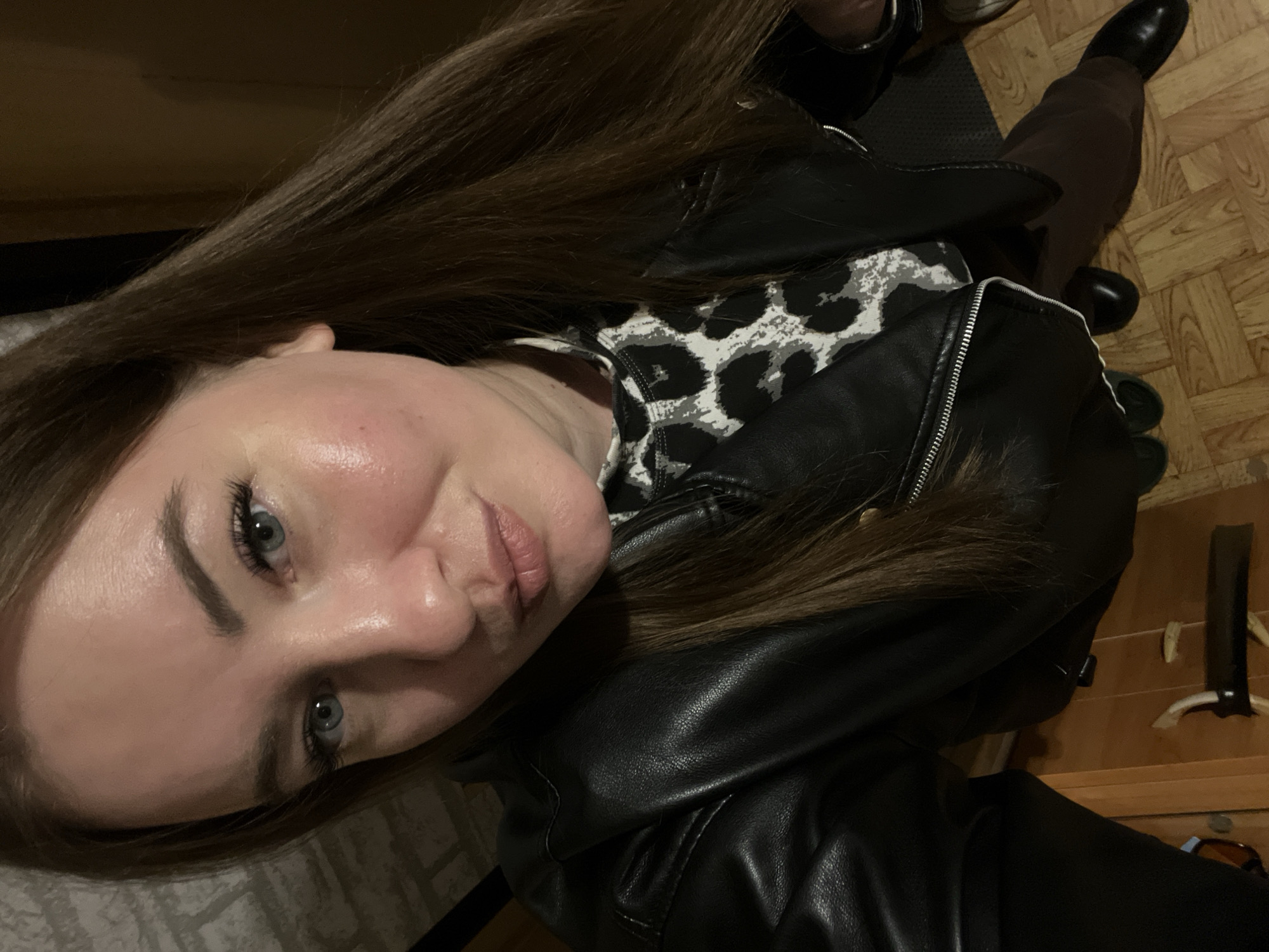 A selfie taken in a location that appears to be indoors, potentially in a residence. The person in the image is wearing a black leather jacket over a patterned top. Part of a black shoe and brown pants are also visible in the background. The floor is wood, and there is a coat rack in the background on a wooden desk. It is not possible to verify if this scene is located in the city of Chaikovsky, Russia.