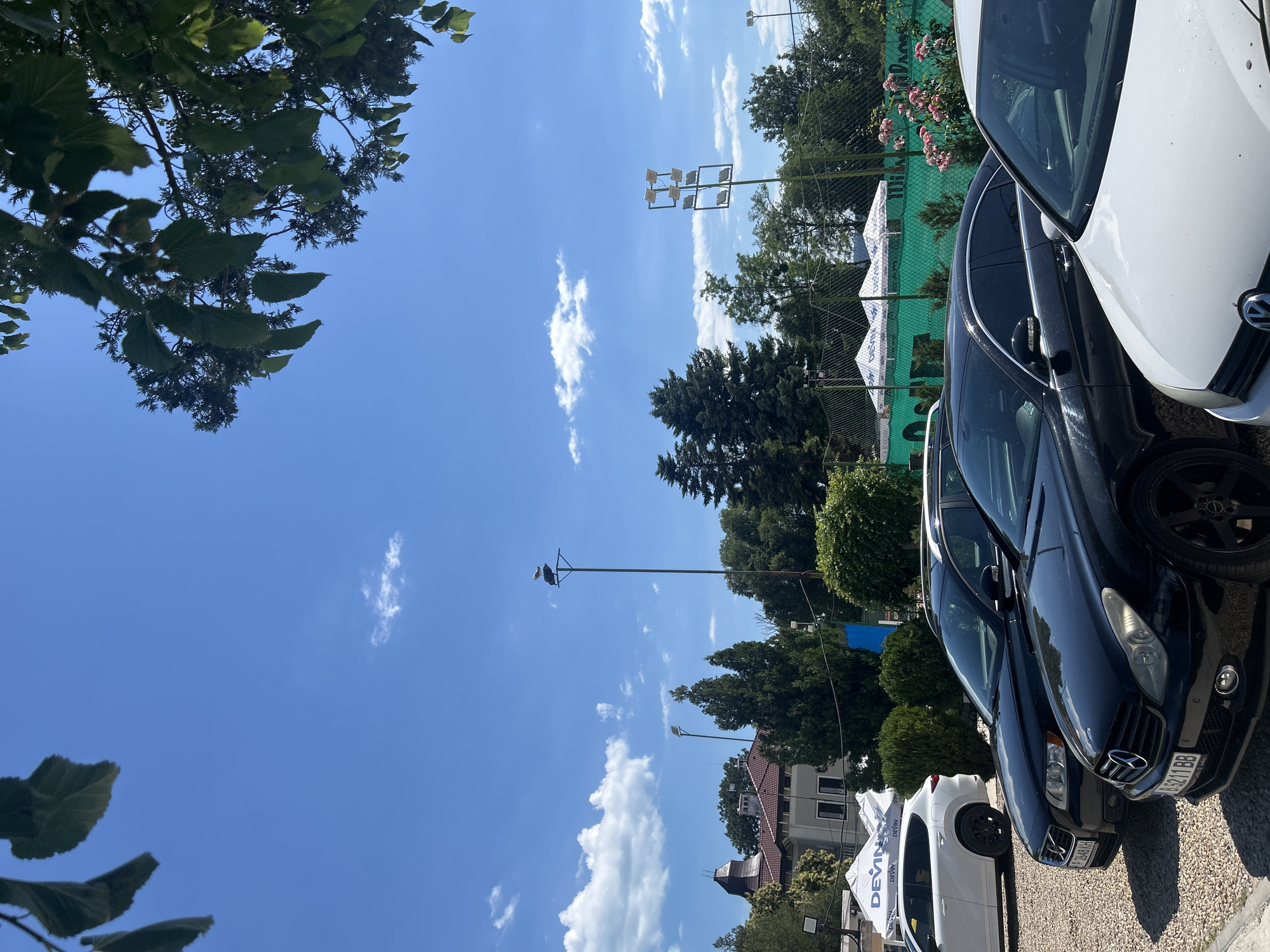This is a medium shot of a parking lot with several cars and a tennis court in the background. The image is taken on a sunny day with a clear blue sky and some white clouds. The trees are green and lush, indicating it is likely summer or late spring.
The primary subjects are the cars parked in the lot. There is a white car in the foreground on the right, partially obscuring a dark-colored car next to it. The dark car has a license plate visible that reads "LZ 621 BB". Further back, more cars are parked, and a white tent with blue lettering is visible. The tent has text that appears to be "DEVIN" repeated several times.
In the background, a green tennis court is visible behind a chain-link fence. There are pink roses growing near the fence. A tall light pole with multiple lights is also prominent in the scene, and on top of another pole, there appears to be a bird's nest with what might be birds. The overall setting suggests a recreational area or sports complex.
The time of day appears to be daytime due to the bright sunlight and clear sky. The weather is fair and sunny. The location is suggested to be Varna, Bulgaria, based on the provided context. There are no people visible in the image, and the emotions conveyed are neutral, reflecting an ordinary outdoor scene.