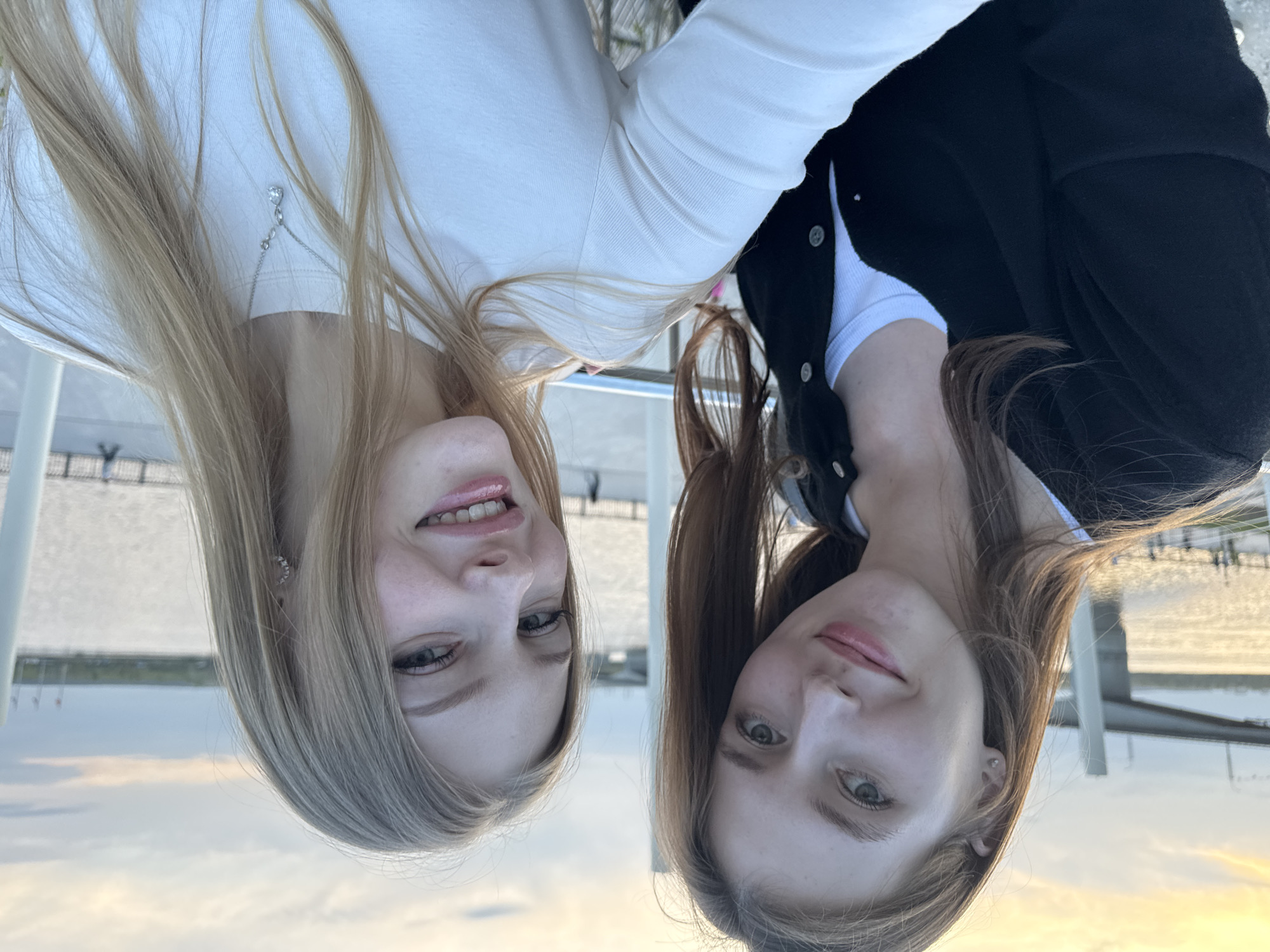 Two young women pose closely together, looking at the camera. The woman on the left has light blonde hair and wears a white long-sleeved shirt. The woman on the right has brown hair and is wearing a black cardigan over a white top. The background shows a body of water and a walkway with railings. Given the presence of water, bridges, and a skyline in the backdrop, the location is likely in Perm, Russia, near the Kama River.