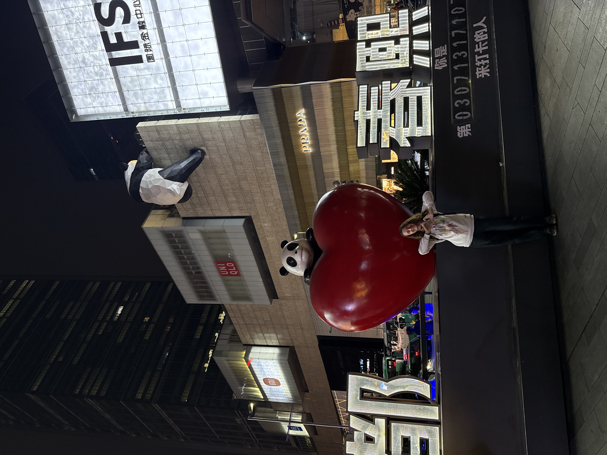 Here is a description of the image:
The photograph shows a night scene on a street in the Jinjiang District of Chengdu, China. A woman stands next to a large, red heart-shaped sculpture that has a panda face attached to it. Behind the sculpture, there's a black counter-like structure with illuminated Chinese characters and numbers "0307131710". The buildings behind the sculpture showcase various brands: "IFS", "PRADA", and "UNIQLO". Another panda sculpture is visible on one of the buildings, appearing as if it's climbing the structure. The surroundings include street lights, other buildings, and hints of traffic lights and vehicles. The overall atmosphere is that of a bustling urban area at night.