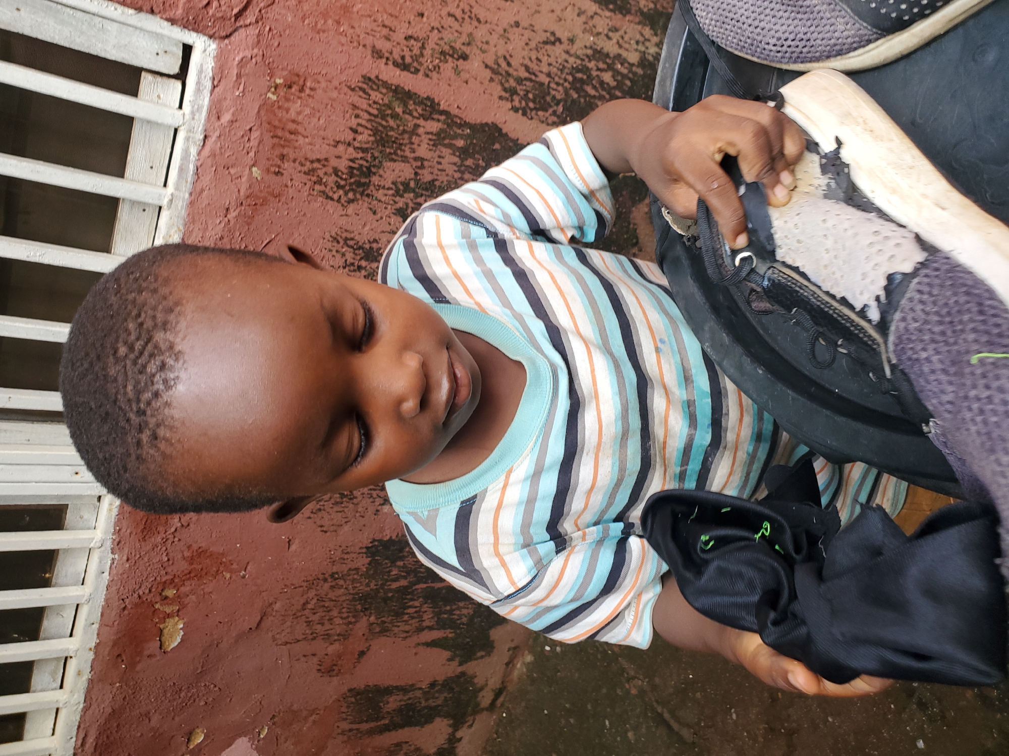 The image shows a young Black child with eyes closed, appearing to be resting or concentrating. The child is wearing a striped t-shirt with horizontal lines of light blue, white, gray, and orange. The child's hands are engaged with a black object that appears to be a shoe, with parts of its sole and interior visible. The child's left hand is holding the shoe open, while the right hand is reaching inside it.
The scene is set outdoors on a reddish-brown, textured surface, likely concrete or asphalt, with some patches of darker discoloration suggesting moisture or dirt. To the left of the child, a white slatted structure, possibly part of a fence or gate, is visible. The lighting suggests it is daytime, and the overall mood is casual and perhaps a bit unkempt, given the condition of the shoe and the ground. The location context provided is Jos, Nigeria. There is no visible text in the image.