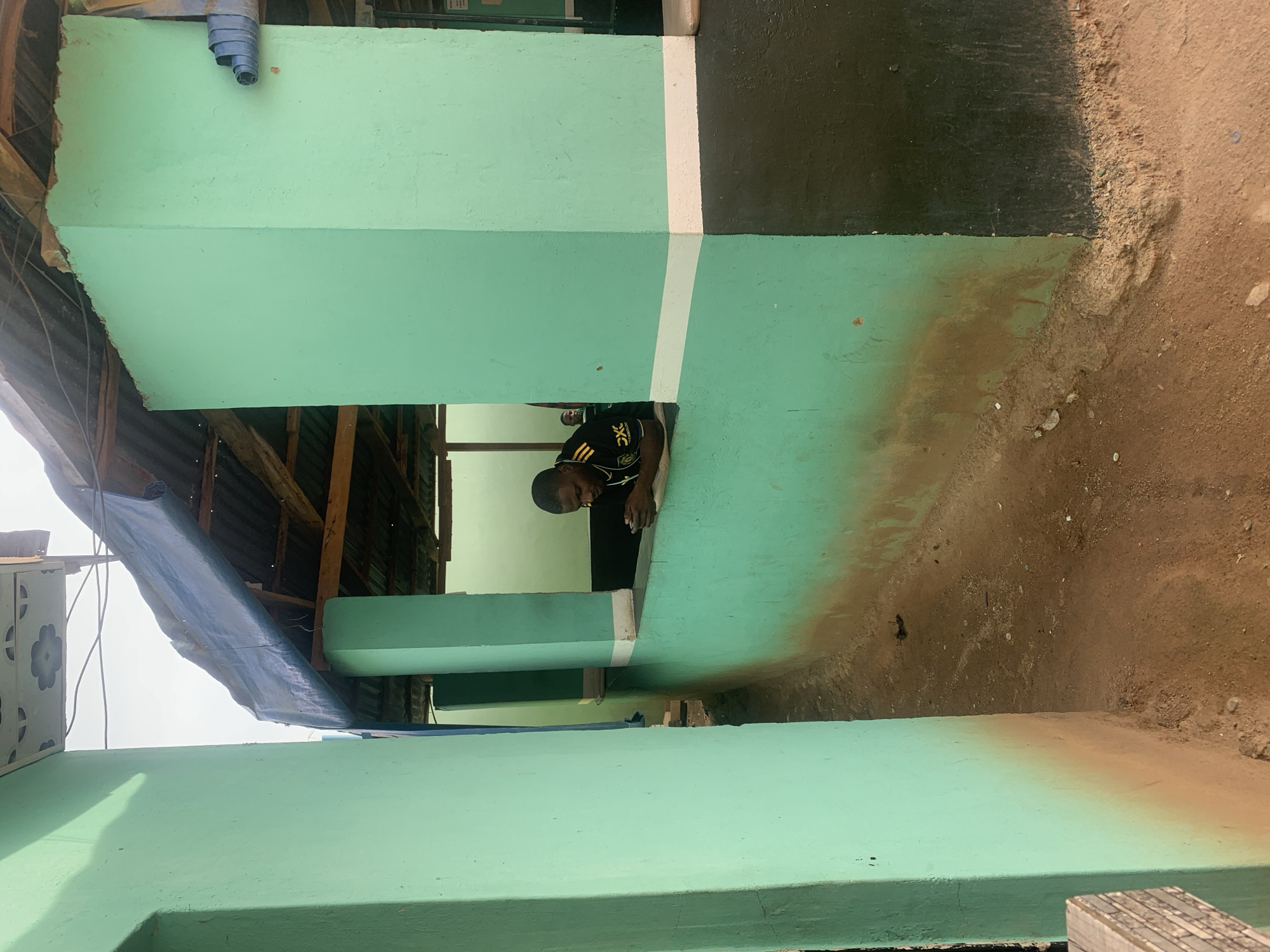 A slightly high-angle, exterior view shows the side of a building in the city of New Stadium, Nigeria. The building is painted a light green color. The building has been added onto to create more space. A man is seen through a partially open section of the building. The ground is brown.