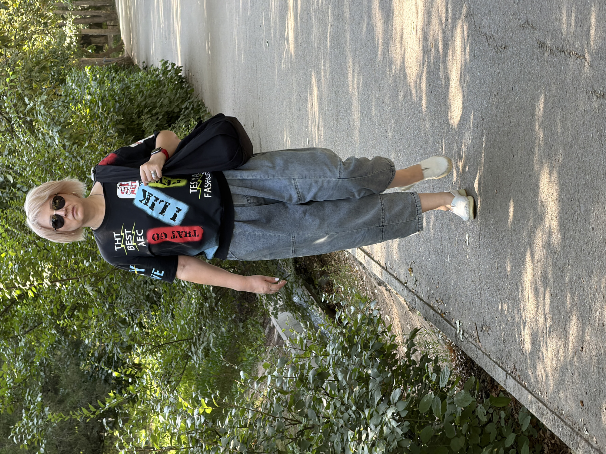 A woman with blonde hair, wearing sunglasses, a black t-shirt with graphic print, loose-fitting grey denim pants, and white slip-on shoes, walks along a paved path in a park. She carries a black shoulder bag. The path is lined with trees on the right and dense green foliage and a concrete drainage ditch on the left. Sunlight casts shadows across the path. The woman is facing the camera and appears to be walking towards the viewer.