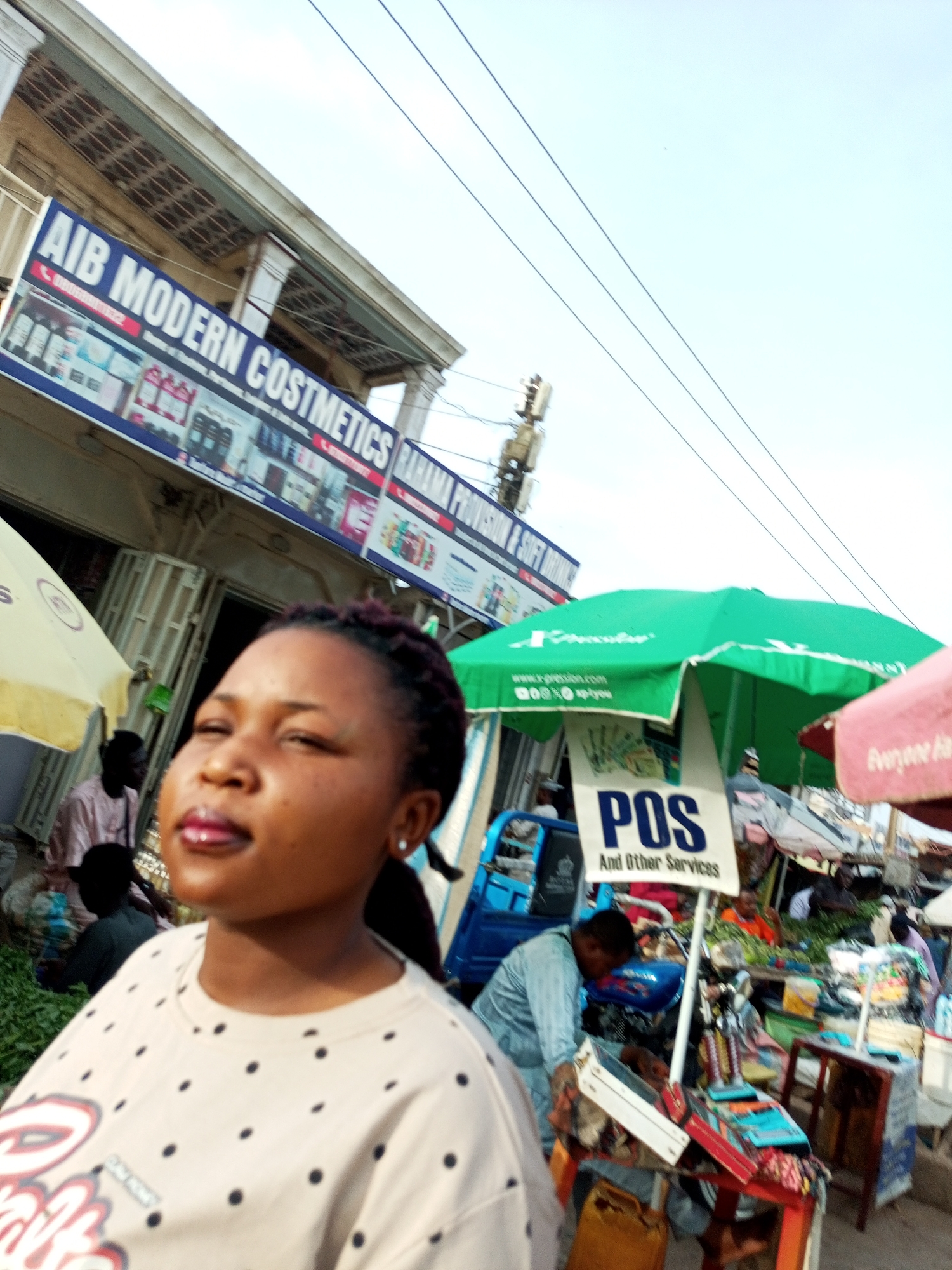 In an outdoor market setting, a woman is in the foreground with her eyes closed and her head tilted back. She is wearing a light-colored t-shirt with a black polka dot pattern. Behind her, a store sign reads "AIB MODERN COSTMETICS RAHAMA PROVISIONS". The market appears to be bustling with activity, featuring various stalls and vendors. A green umbrella with the word "POS" prominently displayed is visible, indicating financial services are available. People are seen browsing and interacting with vendors. The overall scene depicts a vibrant and lively marketplace.