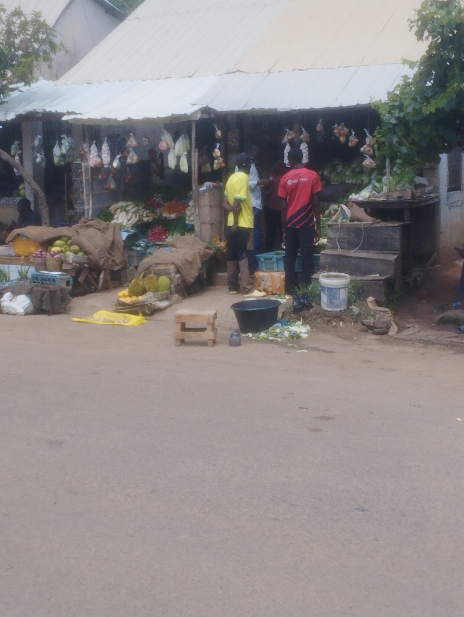The image depicts an outdoor market scene in Jos, Nigeria. In the background, a stall with a corrugated metal roof displays various goods, including produce bagged in plastic and hanging from the awning, and baskets of fruits and vegetables on shelves. Two men are visible in the mid-ground; one, wearing a yellow shirt, stands near the stall, while another, in a red shirt, is facing away from the camera, seemingly interacting with the stall. In the foreground, the market extends onto a paved road. Various items are spread out on the ground, including burlap sacks covering produce, baskets of fruits (possibly oranges and durians), a black bucket, and a small wooden stool. The overall environment suggests a bustling local market.