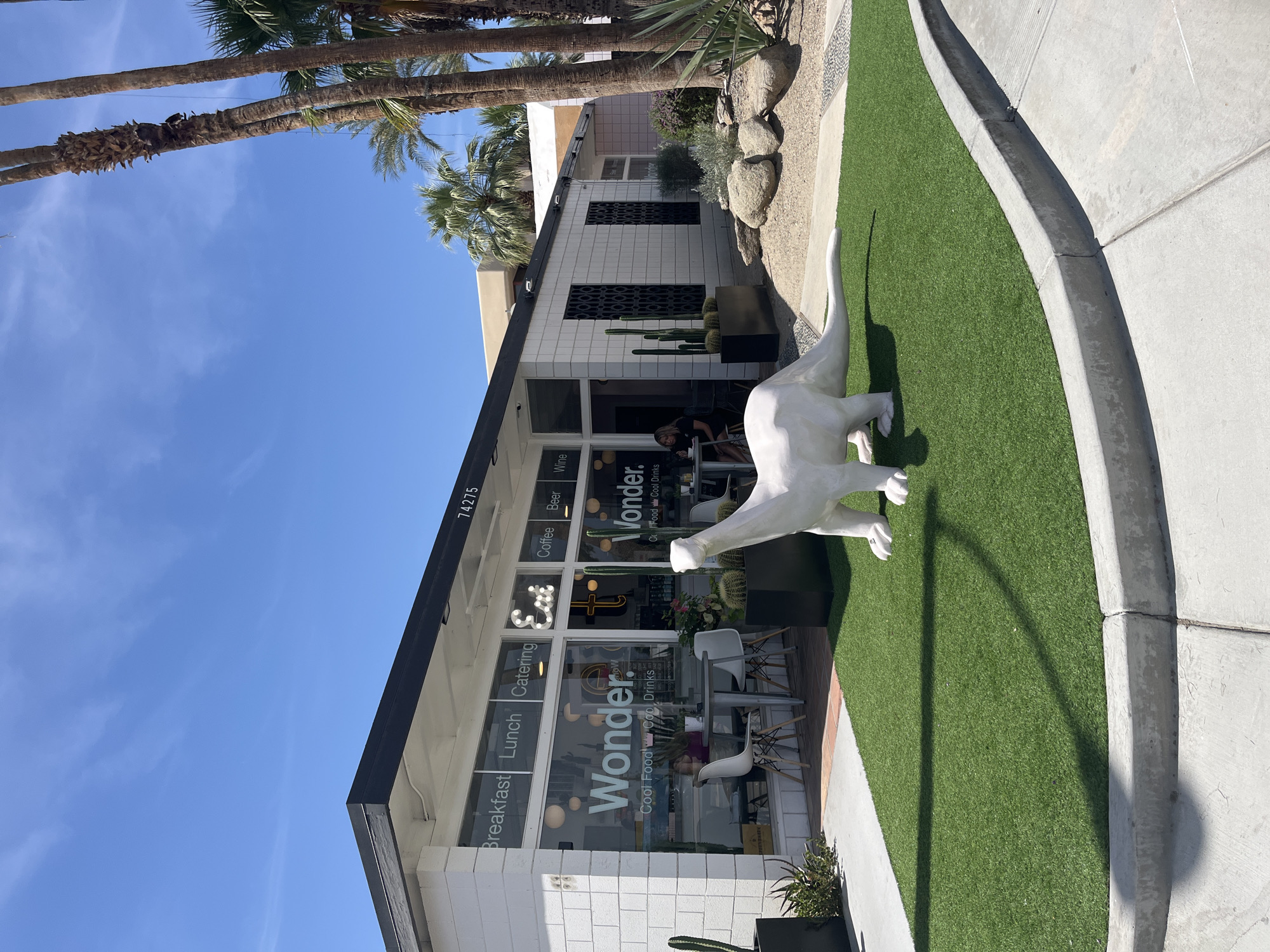 Here is a description of the image:
The photograph depicts the exterior of the "Wonder" restaurant, located at 74275 in Palm Desert, California. The building features white-tiled walls, glass windows displaying the restaurant's name and offerings (Breakfast, Lunch, Catering, Coffee, Beer, Wine, Cool Food Cool Drinks), and a black trim along the roofline. A large, white dinosaur statue stands on a patch of artificial turf in front of the restaurant. Palm trees are visible in the background against a clear blue sky. The surrounding area includes a concrete sidewalk and landscaping with rocks and plants. People are seated inside the restaurant.