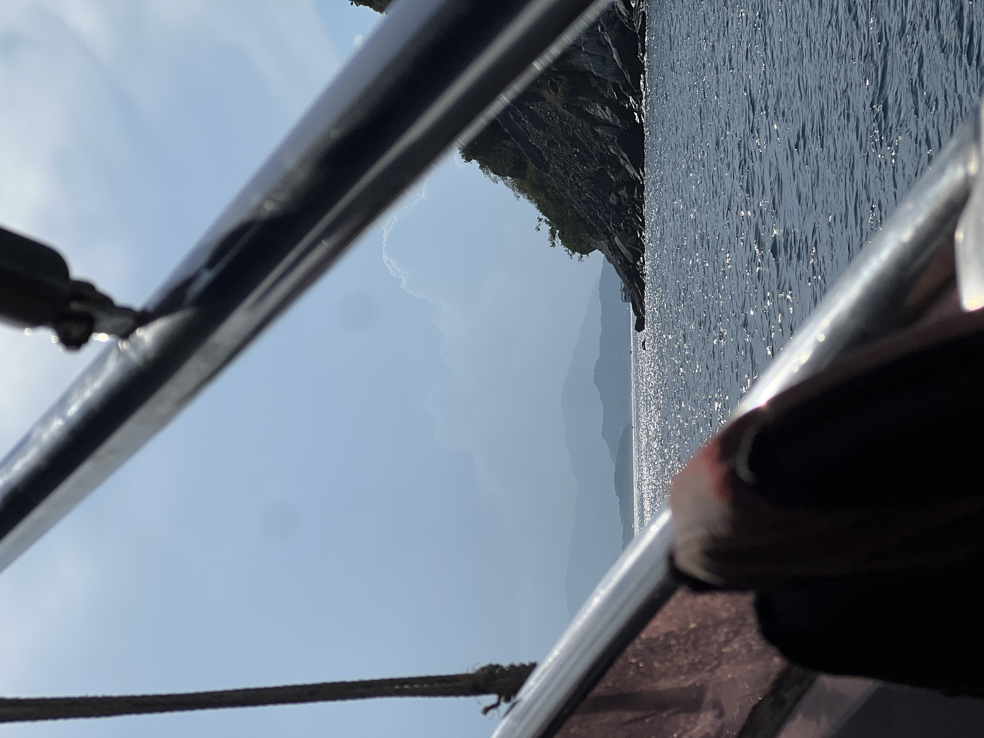 The image, taken from a low angle perspective, shows a seascape viewed through the railings of a boat. The sea is visible, reflecting sunlight. A rocky coastline is partially visible. Beyond the coastline are distant mountains. The sky is partly cloudy. The railings of the boat, made of metal, are in the foreground and appear to be wet, as indicated by water droplets. A rope is also partially visible in the foreground.
It is not possible to confirm that this image was taken in Bac Nha Trang Ward, Vietnam based on the image alone.