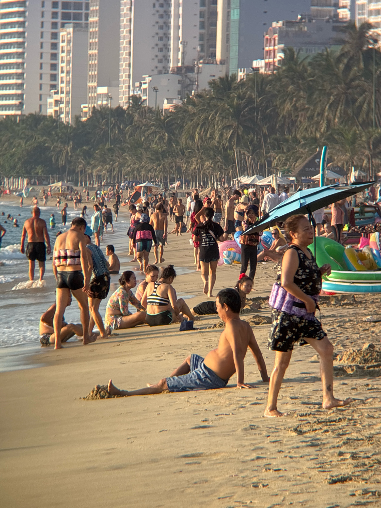 This photograph captures a bustling scene on a sandy beach in Nha Trang, Vietnam, with the city's prominent skyline of tall buildings and palm trees in the background. The beach is populated with numerous individuals enjoying the ocean and the sun. In the foreground, several people are visible on the sand. A man in blue swim trunks reclines on the beach, propped up by his hands, while another man, also in blue swim trunks, sits further back. To the right, a woman with dark hair, wearing a black and white patterned sleeveless top and black shorts, walks across the sand, holding a purple bag. She is partially shaded by a large blue umbrella held by another person.
In the mid-ground, more beachgoers are present, some wading in the shallow ocean waves. A man wearing black swim trunks stands near the water's edge, facing away from the camera. Beside him, another man in black swim trunks, with what appear to be flotation devices strapped to his back, walks forward. Several other individuals are seen scattered across the beach, including a woman in a black top and floral print shorts, sitting on the sand with another person. Further back, a dense line of palm trees separates the beach from the urban landscape, where numerous multi-story buildings dominate the horizon. The overall impression is one of a lively, sunny day at a popular tourist destination.