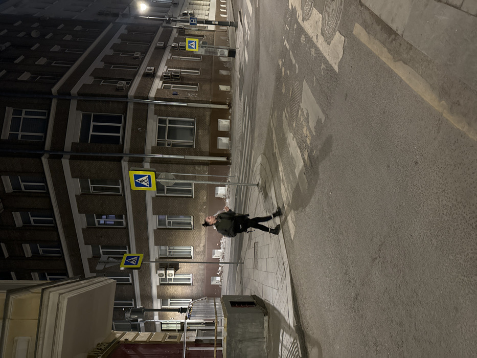 The image depicts a nighttime street scene at the intersection of Bolshoy Ovchinnikovsky Pereulok and Sredny Ovchinnikovsky Pereulok in Moscow, Russia. The prominent building on the corner, which appears to be 10 Bolshoy Ovchinnikovsky Pereulok, is a multi-story (at least five visible floors) brick structure with a dark brown facade and numerous white-framed windows, several of which have exterior air conditioning units.
A single female subject, dressed in a dark jacket, dark pants, and dark boots, is observed walking from left to right across a faded pedestrian crossing. Her hair is pulled back into a bun. Her head is turned slightly to the left, and her mouth is partially open, possibly in mid-speech or exhalation. She holds a small, light-colored object in her right hand.
The street is an asphalt road with visible wear, featuring faded white zebra-stripe pedestrian crossing markings and several circular manhole covers. A yellow line is painted along the right curb. The sidewalks are paved with lighter-colored square tiles.
Multiple poles are present. Two prominent poles, positioned on the left side of the intersection, display identical square blue pedestrian crossing signs, each featuring a white triangle enclosing a black pedestrian icon. On the right side, a pole supports a street lamp and a blue rectangular street sign indicating "Б. Овчинниковский пер." (Bolshoy Ovchinnikovsky Pereulok) and "Ср. Овчинниковский пер." (Sredny Ovchinnikovsky Pereulok). Further down the street on the right, another blue "P" sign, indicating parking, is visible.
On the immediate left, a portion of another building features a light-colored upper section and a darker red lower section with a visible red door. A concrete structure with metal railings is also present on the left corner, adjacent to the sidewalk. The scene is illuminated by artificial streetlights, casting shadows from the poles and the walking subject.
