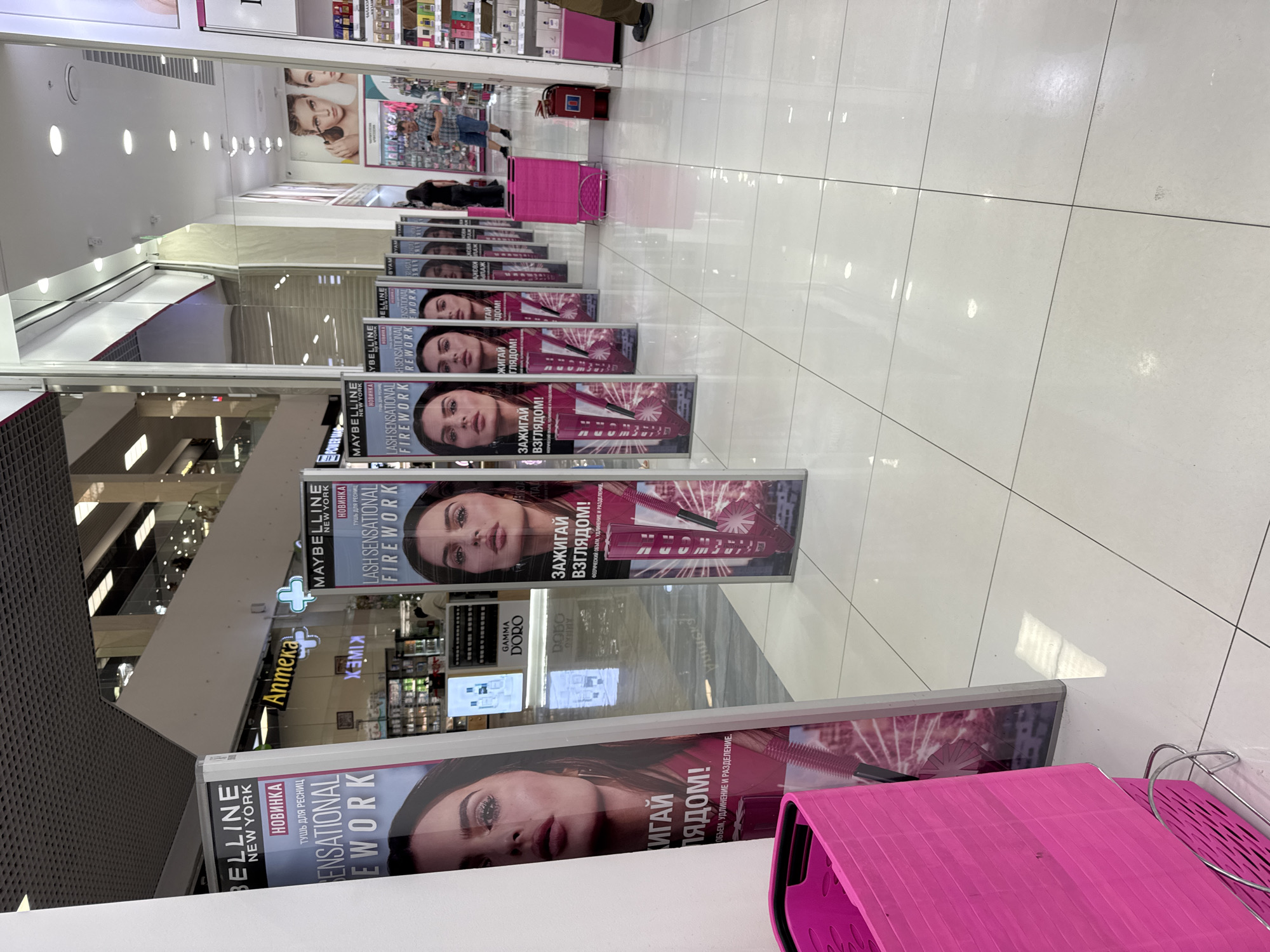 The image depicts the interior of a retail establishment, possibly a cosmetics store, with a focus on a display of Maybelline mascara advertisements. Multiple vertical banners, each featuring a close-up of a woman's face and product imagery, are arranged in a row. A bright pink shopping basket is prominently positioned in the foreground on the left. The floor is tiled with large, light-colored squares. In the background, a person is visible walking, and shelves stocked with products line the walls. The store appears to be well-lit with overhead lighting. The text on the advertisements is in Russian, indicating the location may be within a Russian-speaking region. While the image does not explicitly state it is in Oskemen, Kazakhstan, it is a plausible location given the presence of Russian language in retail environments within Kazakhstan.