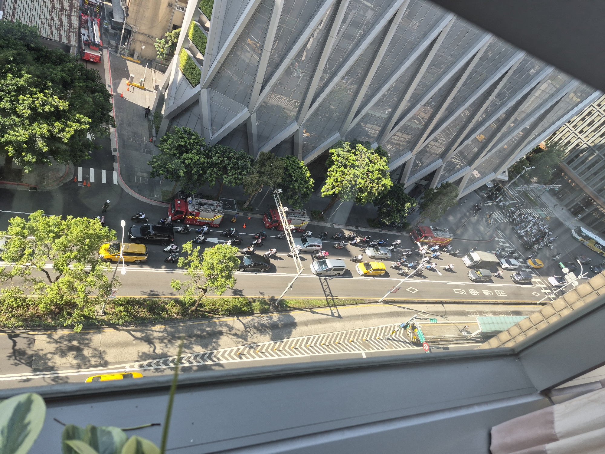 This is an aerial, eye-level view taken from a window overlooking a city street in Taipei, Taiwan. The scene is dominated by a multi-lane road with vehicular traffic and a modern building with a distinctive angular, glass facade.
On the left side of the street, a red fire truck is visible parked. Further down the road, more emergency vehicles, including what appears to be another fire truck and an ambulance, are present. Several individuals, likely emergency responders, are seen near these vehicles. Traffic on the road includes numerous motorcycles, cars, and yellow taxis. Trees line the side of the road, some with lush green foliage.
The modern building on the right features a repeating pattern of angled glass panels, creating a textured and reflective surface. The street itself is marked with white lines and pedestrian crossings. Shadows from the trees and buildings are cast across the asphalt. A few people are visible walking on the sidewalks and crossing the street. The overall impression is of an active urban environment, possibly with a scene of emergency response or a traffic incident.
