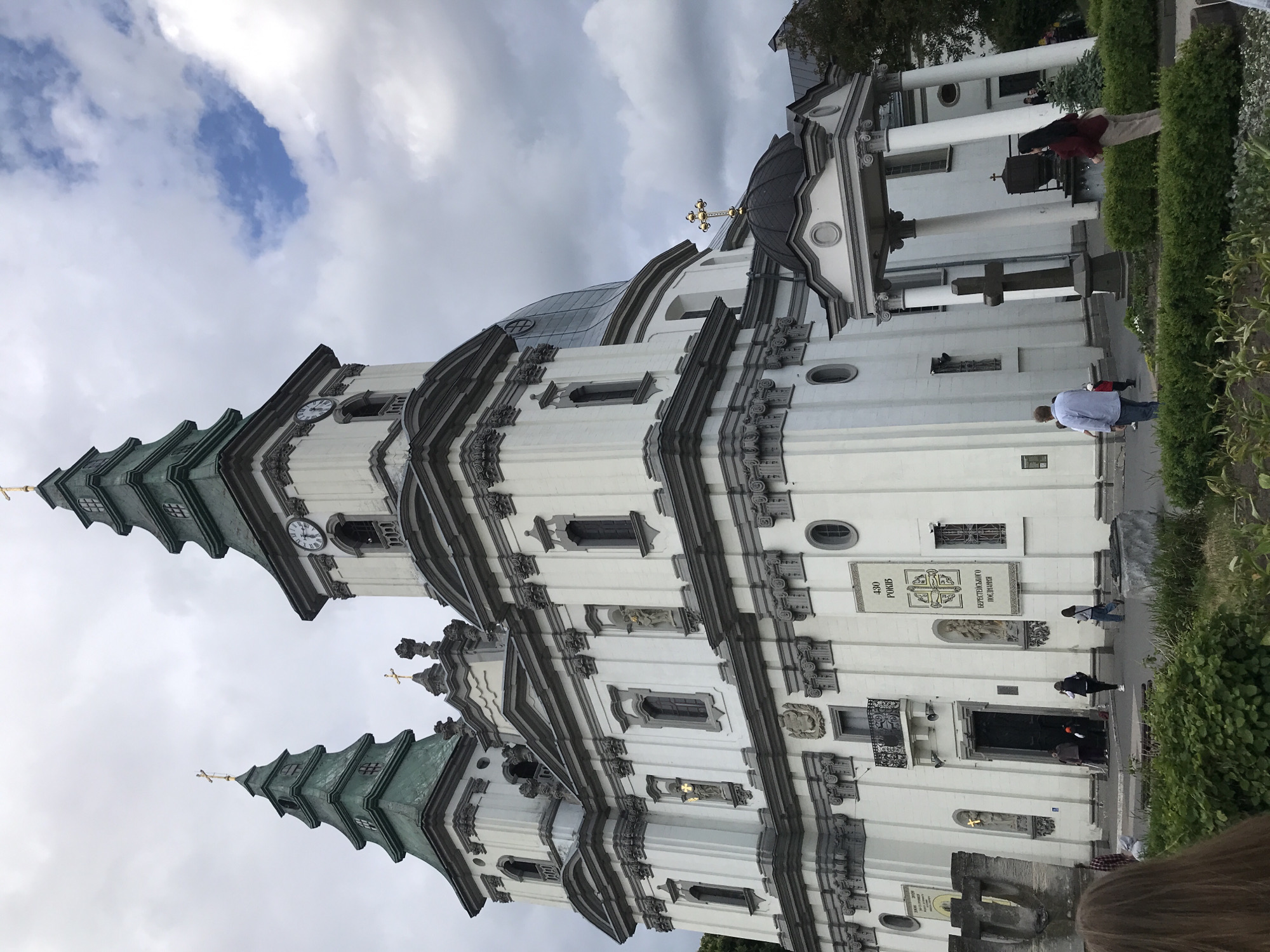 The image shows the Ternopil Archcathedral, a prominent baroque church in Ternopil, Ukraine. The building is mostly white with dark gray trim and is composed of multiple levels, topped with green tiered spires and golden crosses. A large clock face is visible on one of the towers. People can be seen walking around the church at ground level. The sky is overcast with patches of blue. Green shrubbery is visible along the base of the church and in the foreground.