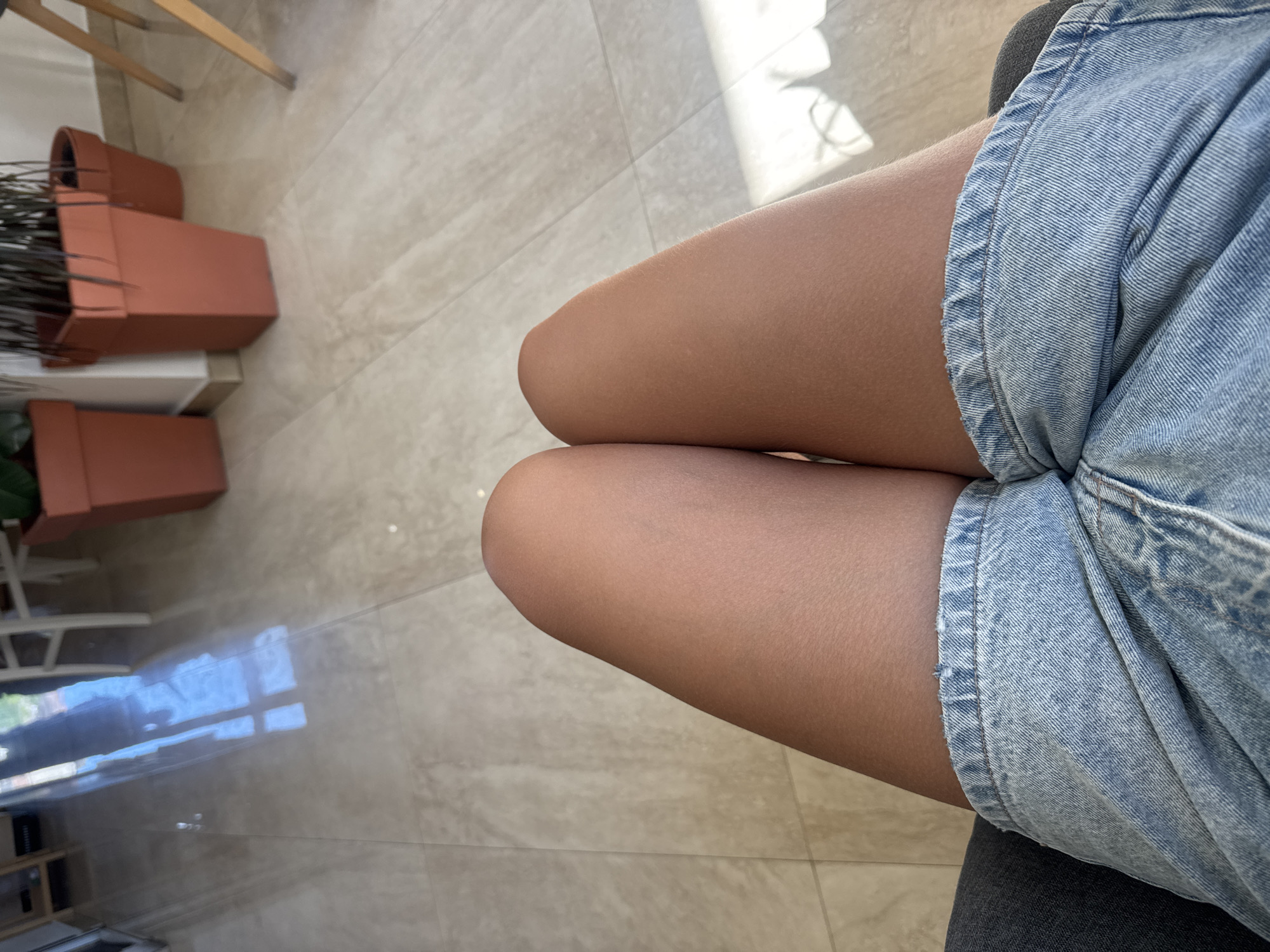 The image shows a close-up view of a person's legs wearing denim shorts, seated indoors on what appears to be a couch or chair. The person's bare legs are visible from the knees down. The environment includes a tiled floor with a sheen, reflecting sunlight coming from a window or doorway. In the background, there are terracotta planters containing plants and parts of furniture, suggesting an indoor space like a cafe, restaurant, or lobby. The location is identified as the city of Budva, Montenegro.