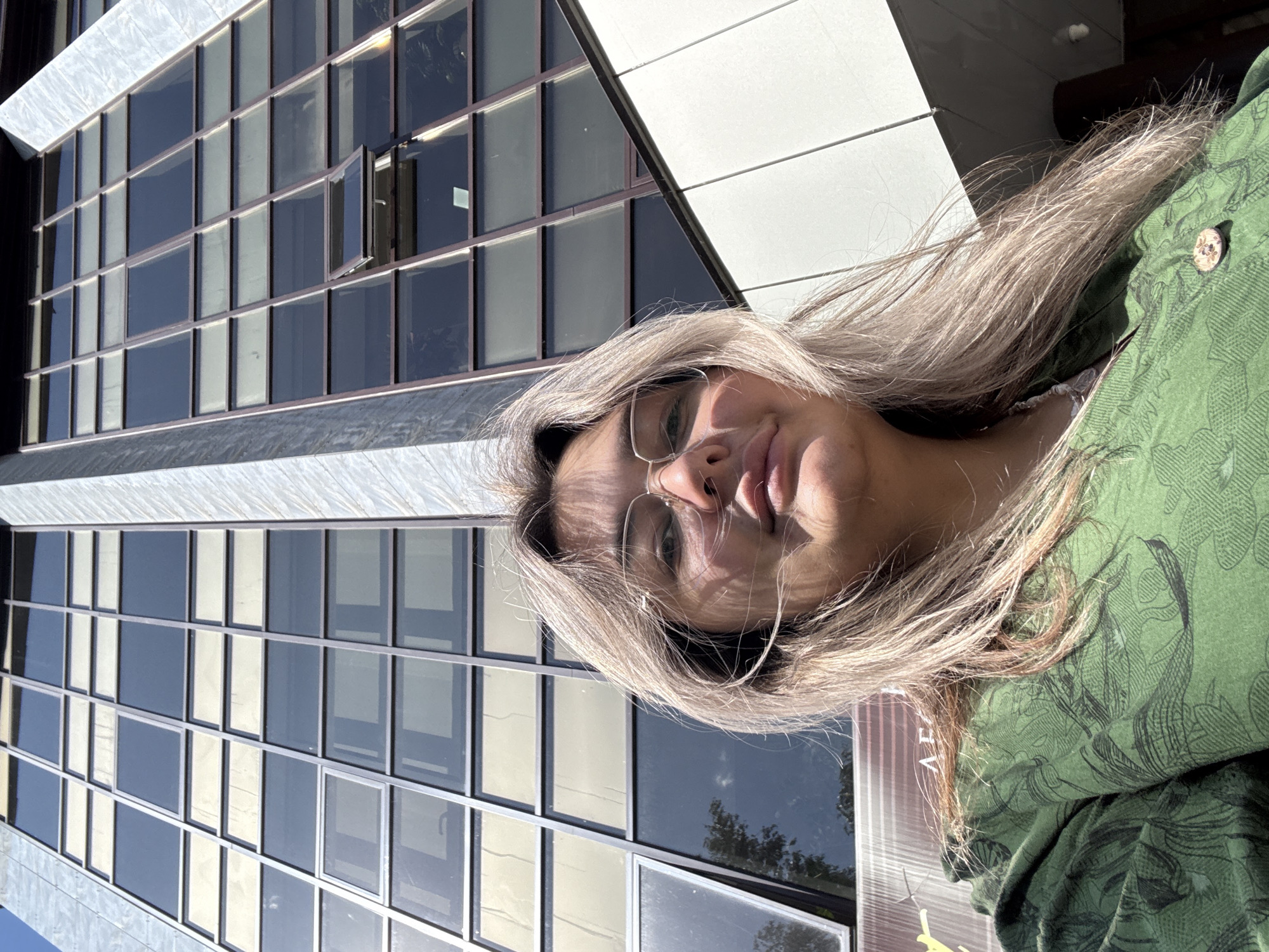 A young woman with blonde hair and glasses is posing for a selfie in front of a modern building. The building features a facade of large, rectangular windows set in a grid pattern. One of the windows is slightly ajar. The woman is wearing a green patterned shirt. The background suggests an urban environment, likely in Birobidzhan, Russia, given the context. The lighting indicates it is daytime, with sunlight casting shadows.